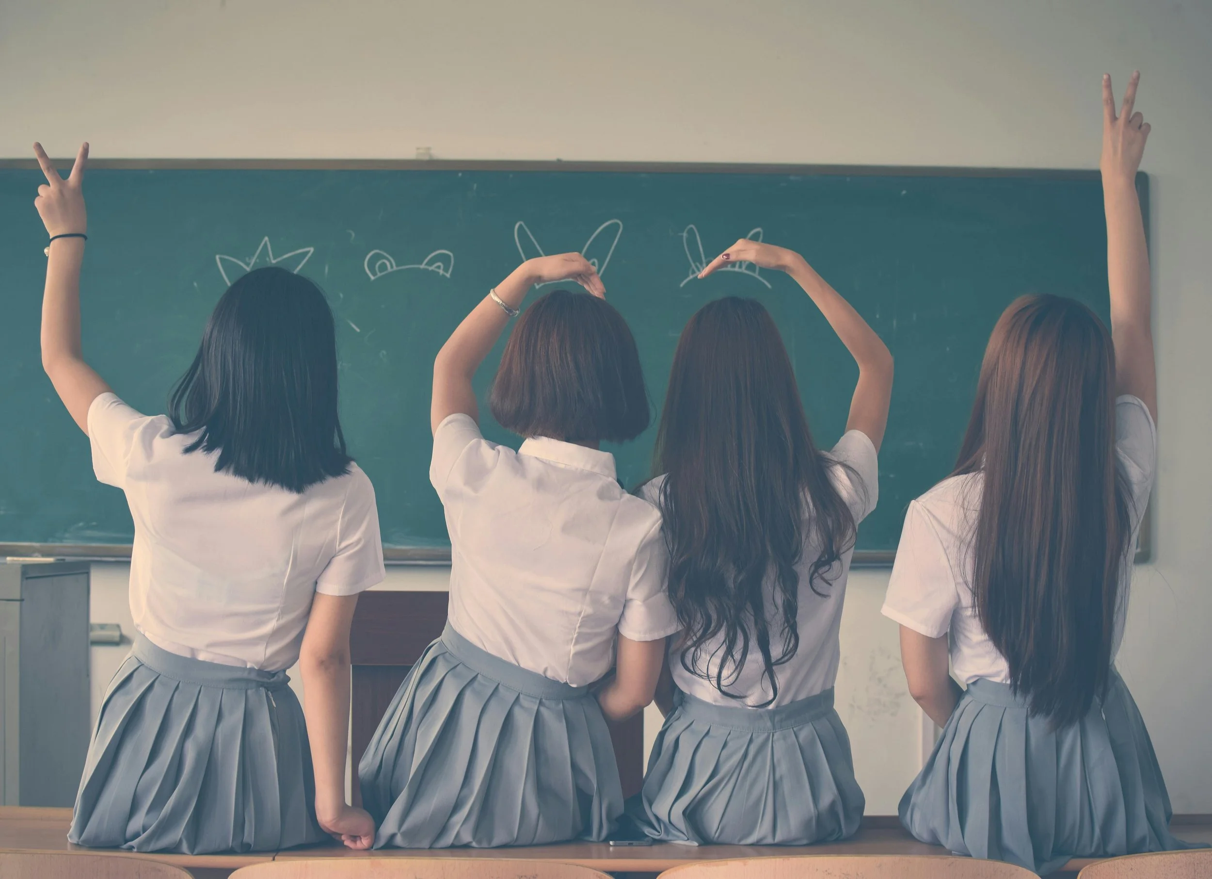 Four female students in school uniforms standing in front of a classroom chalkboard, facing away, with various hand gestures.