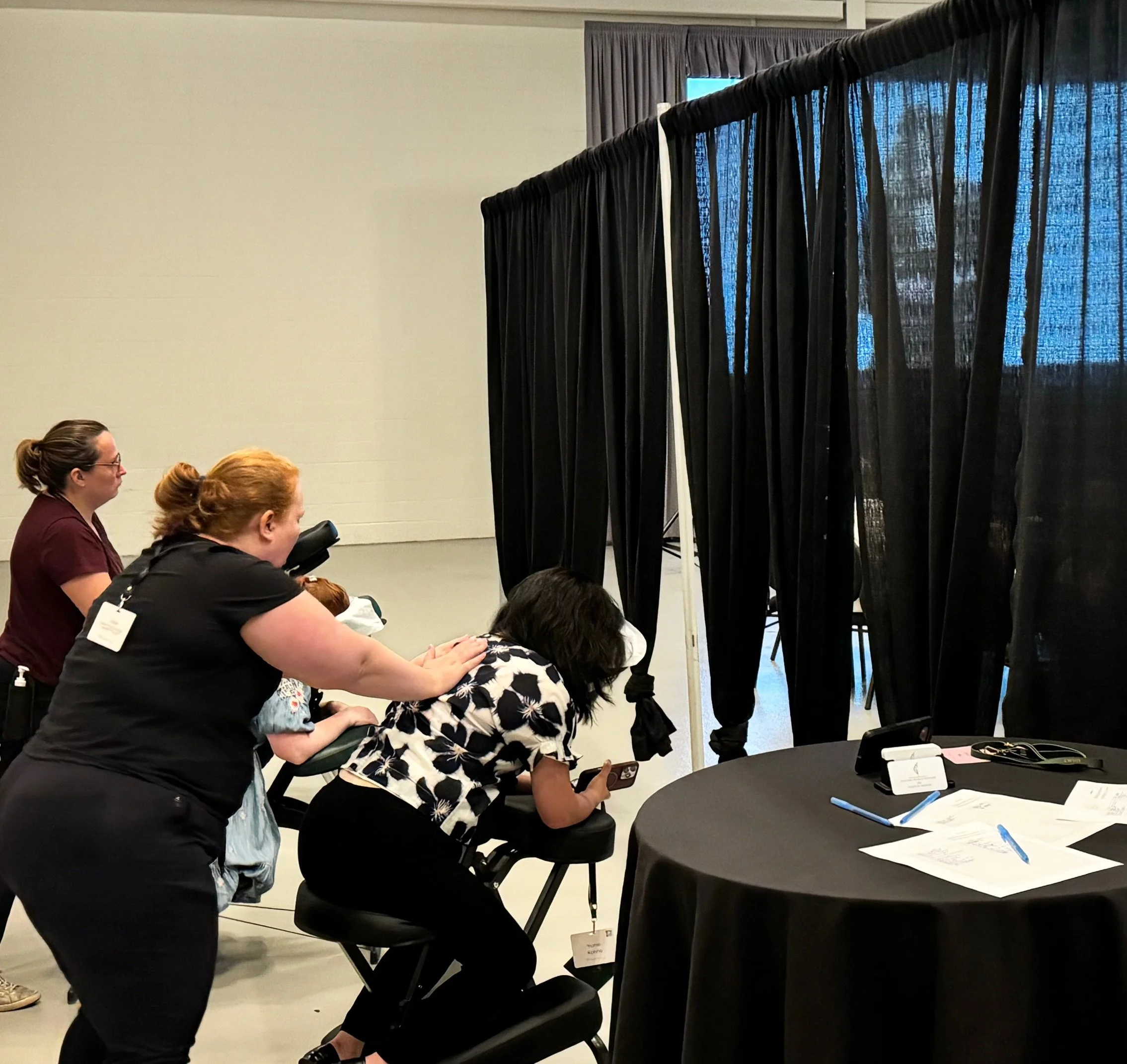 A woman dressed in a floral shirt is seated receiving a massage from a masseuse. Two women are standing nearby, observing. The scene appears to be at a professional or conference setting with a table holding papers and a pen.