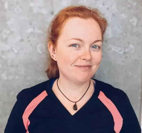 A woman with red hair and blue eyes smiling, wearing a dark top with pink accents and a black necklace, standing against a gray textured wall.