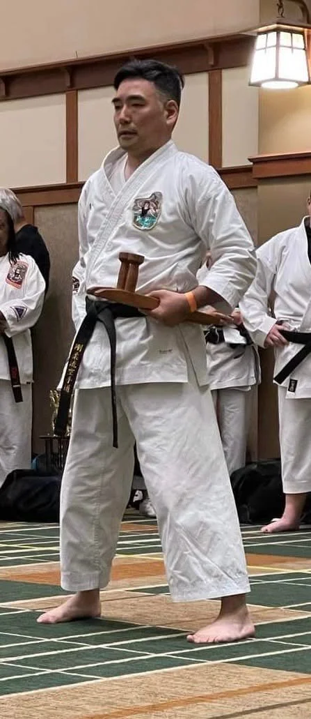 A man in a white martial arts uniform standing in a dojo holding a wooden board and a tattered post, surrounded by other martial artists in similar uniforms.