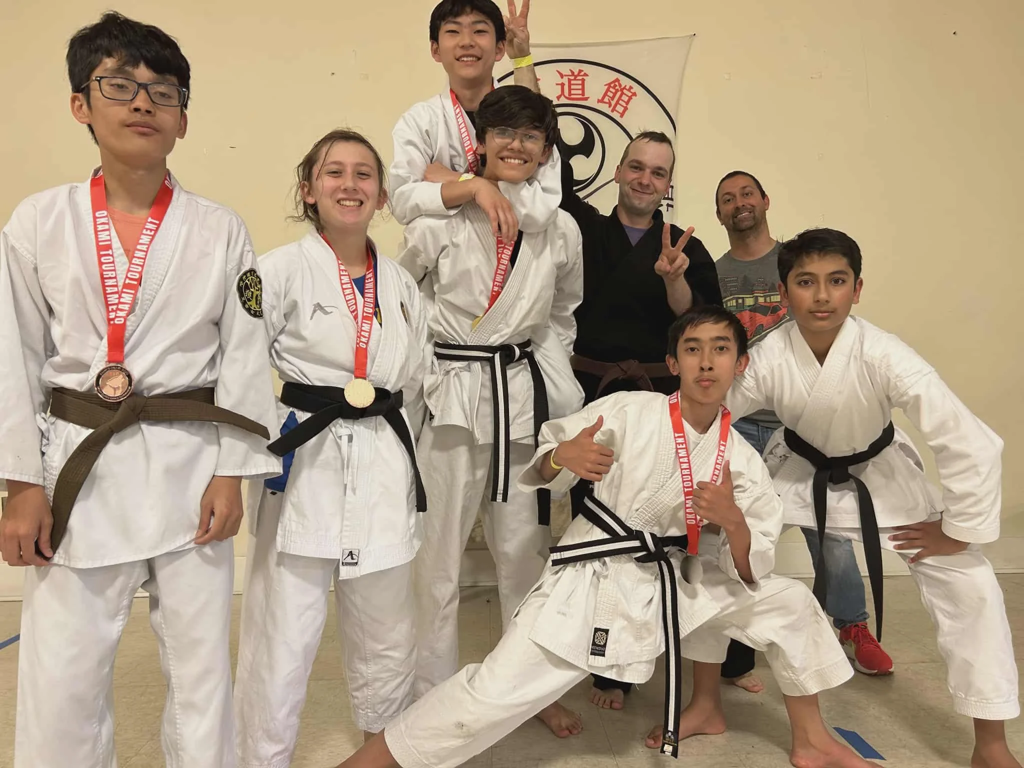 Group of young martial artists in judo uniforms with medals, smiling and posing indoors, with two adults in the background making peace signs and playful gestures.