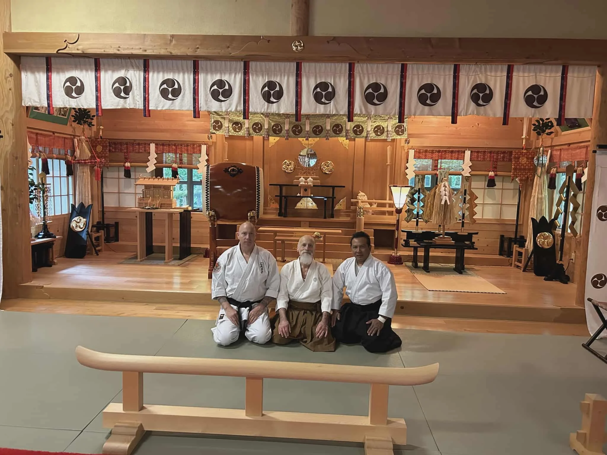 Three martial artists in traditional uniforms kneeling on a dojo mat in front of a wooden altar with various objects, inside a wooden room decorated with Japanese ornaments and symbols.