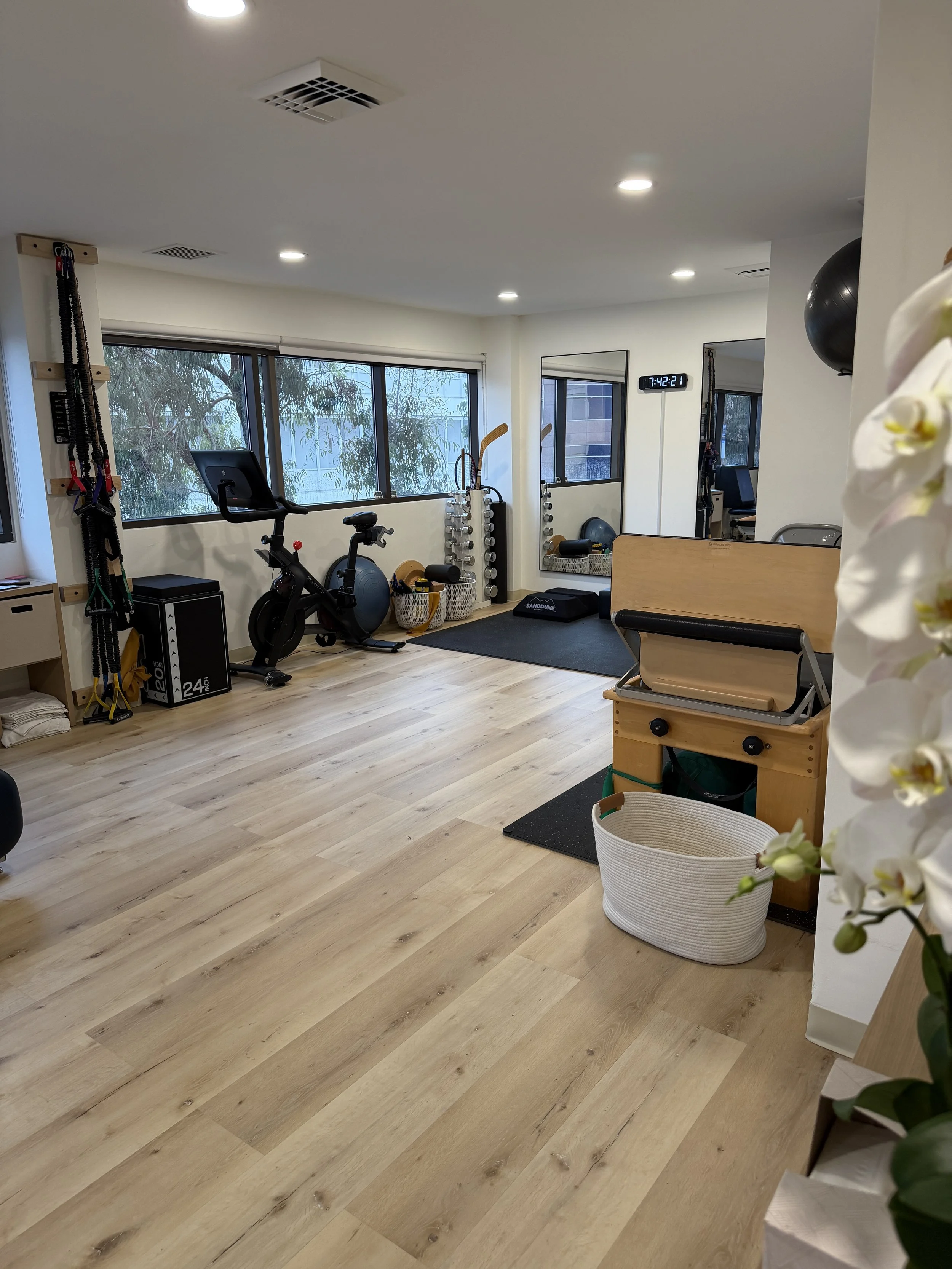 A clean home gym with hardwood floors, large windows, exercise equipment including a stationary bike and a medicine ball, and a wall clock showing 7:42.