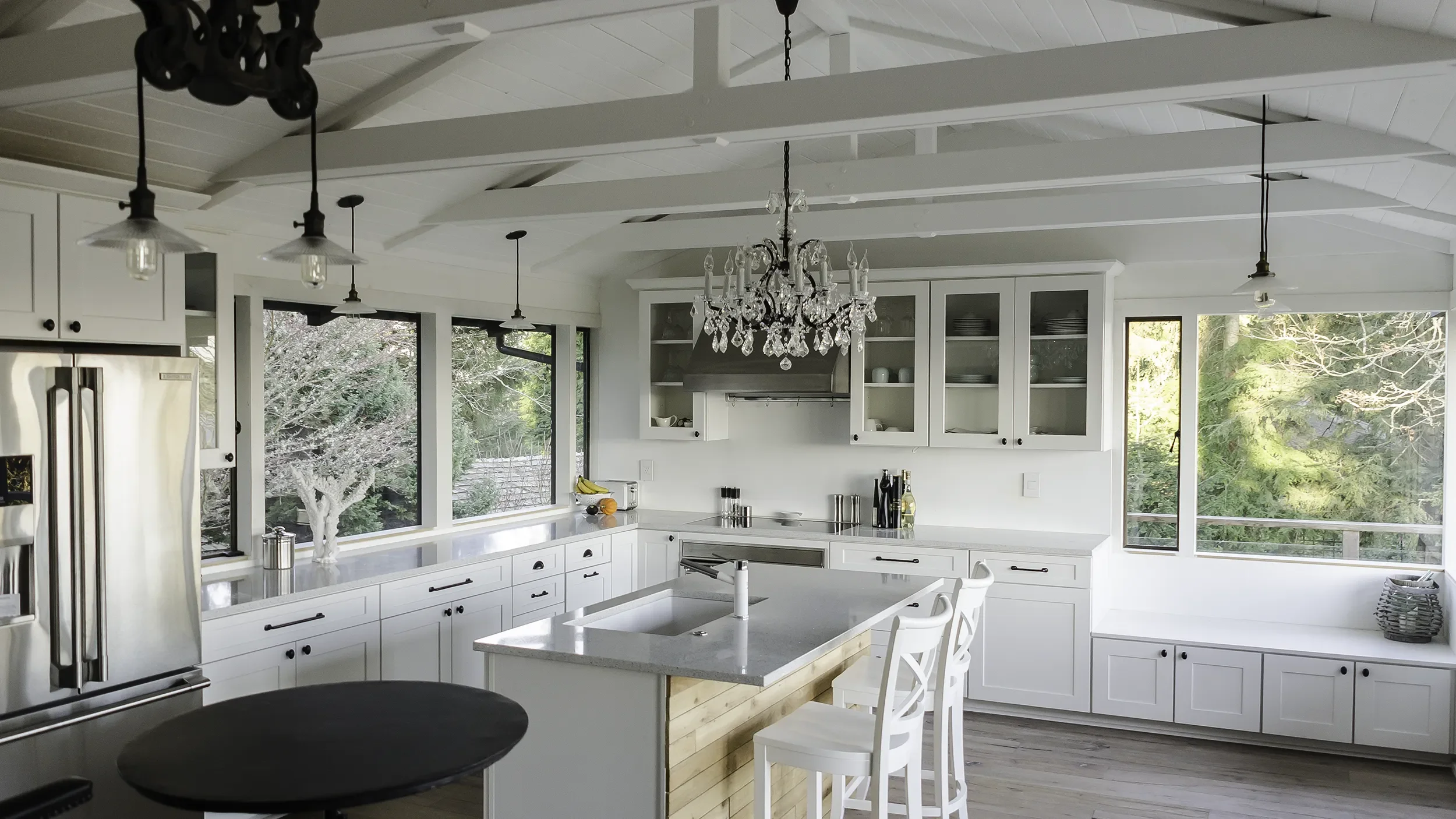 Bright white kitchen with large island, chandelier, stainless steel refrigerator, open cabinets, and large windows with a view of trees outside.