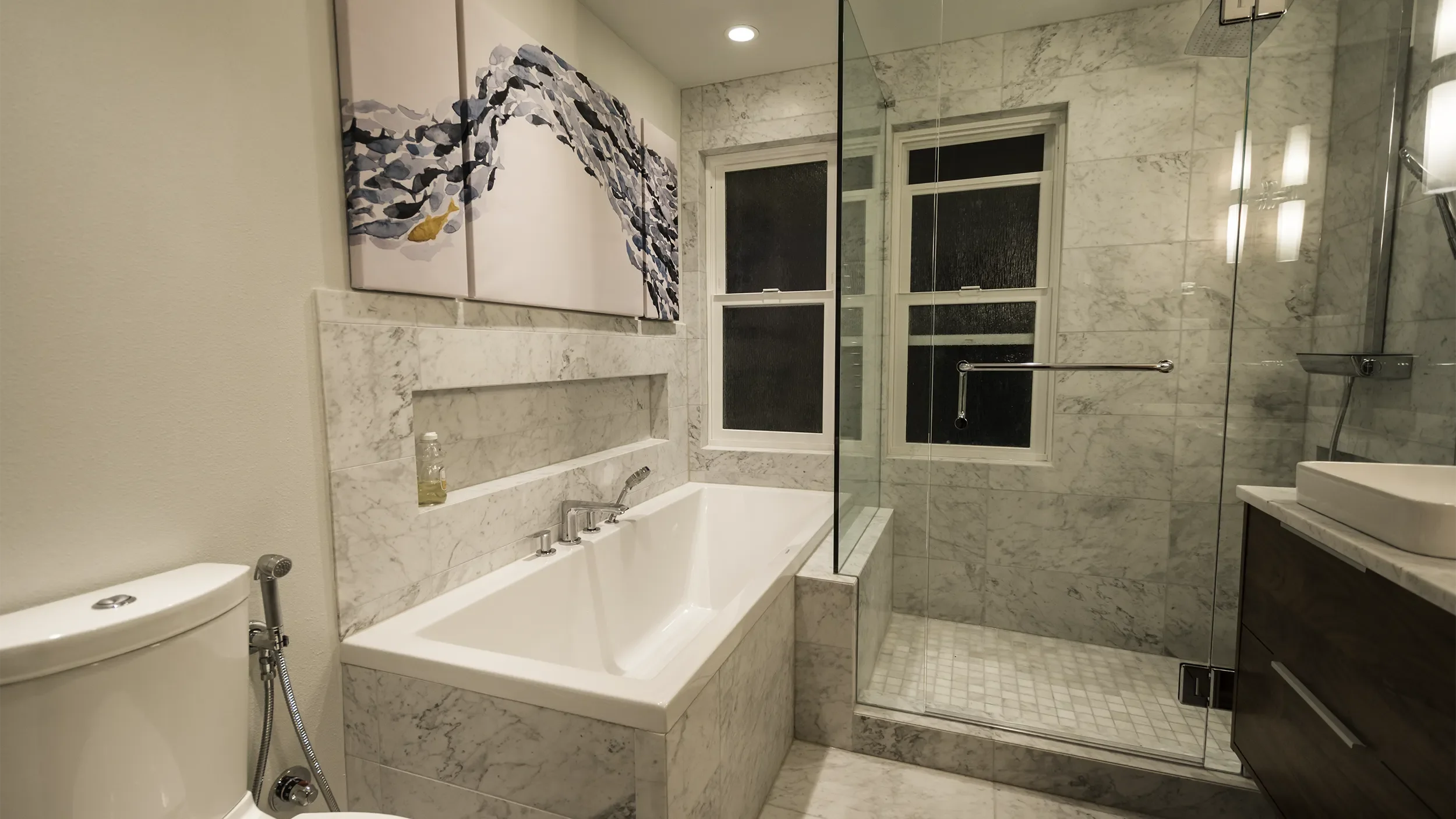 A modern bathroom featuring a bathtub with marble surround, a glass-enclosed shower, a window, a vanity with a sink, and artwork on the wall.