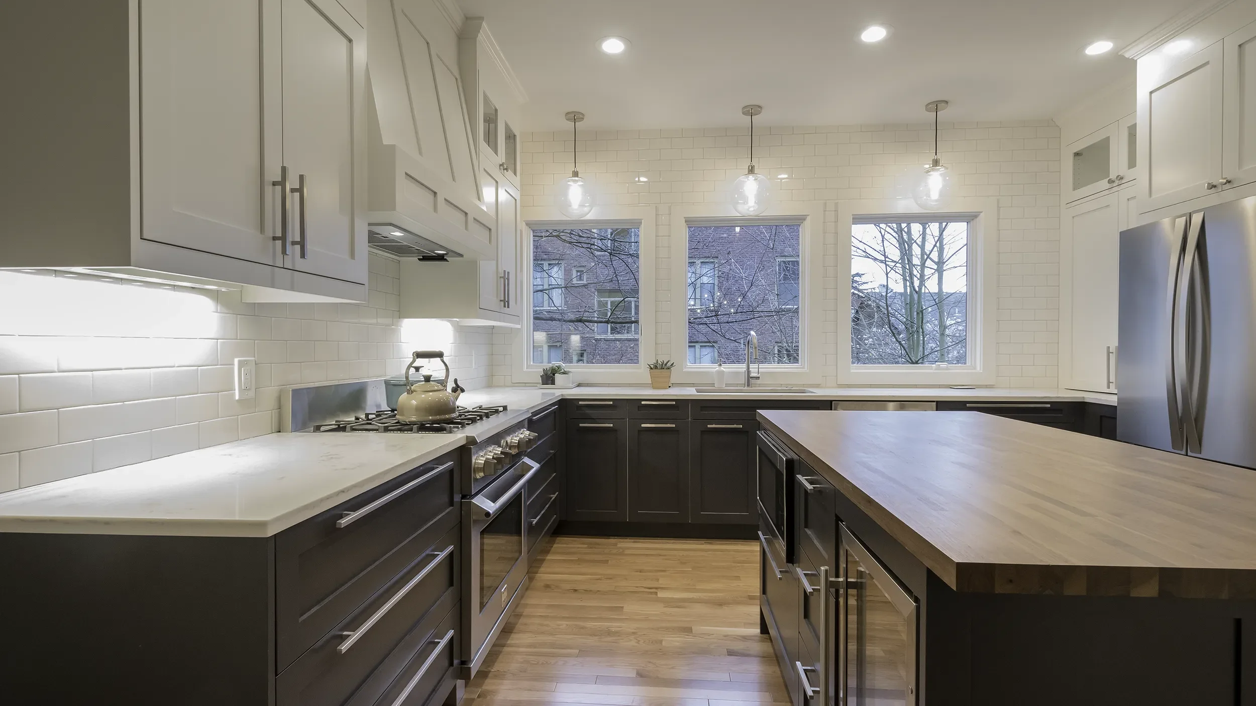 Modern kitchen with white upper cabinets, black lower cabinets, a large wooden island, stainless steel appliances, white subway tile backsplash, three pendant lights, and three windows with trees outside.