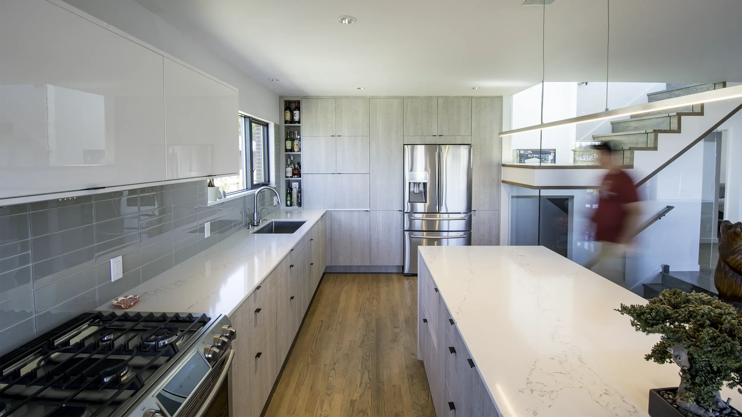 Modern kitchen with light wood cabinets, white countertops, stainless steel refrigerator, gray tile backsplash, and wood flooring. A man in a red shirt is walking near stairs with glass railing. A small potted bonsai tree is on the island.
