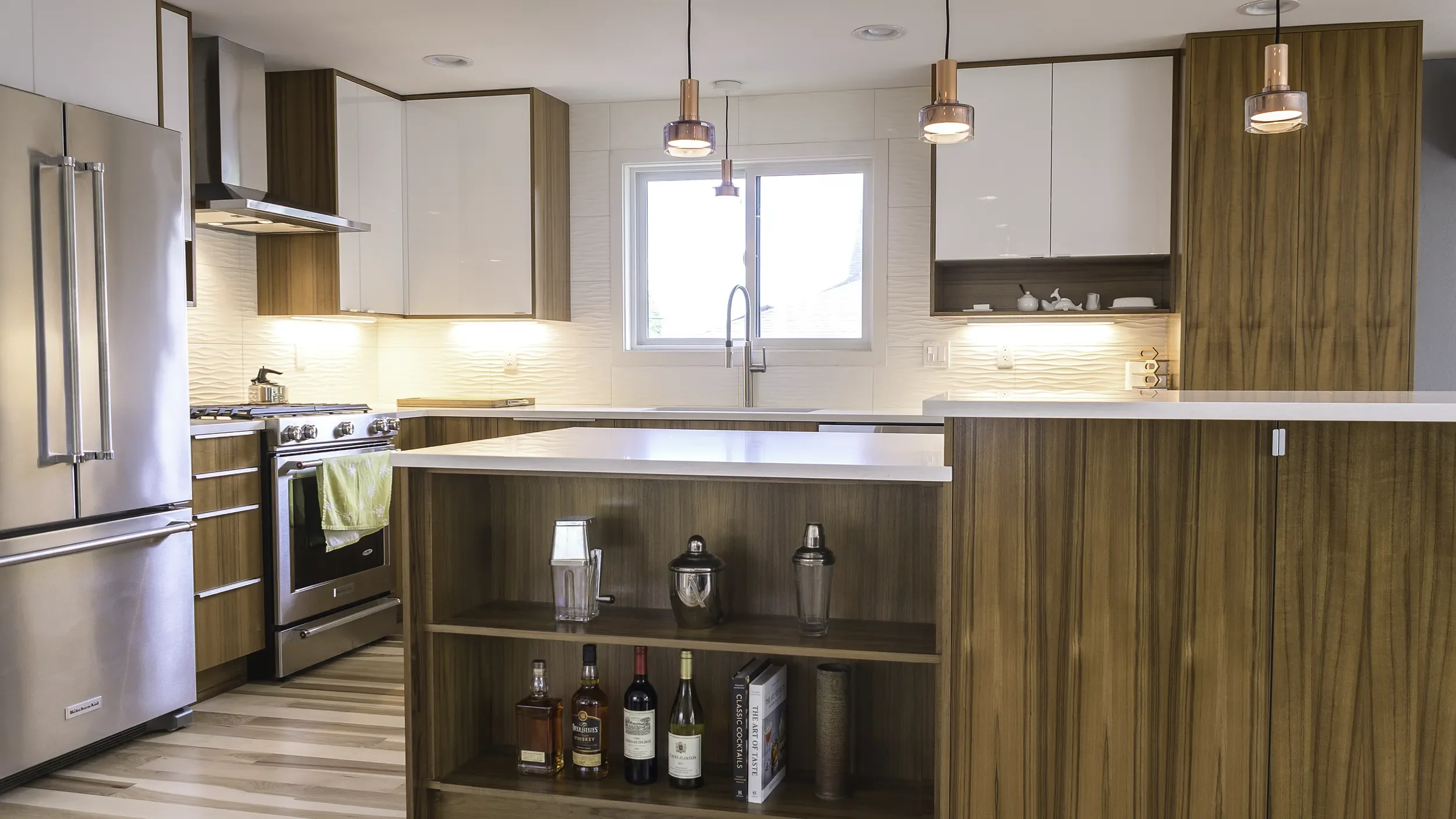 Modern kitchen with wood and white cabinets, stainless steel refrigerator and oven, hanging pendant lights, and a window above the sink.