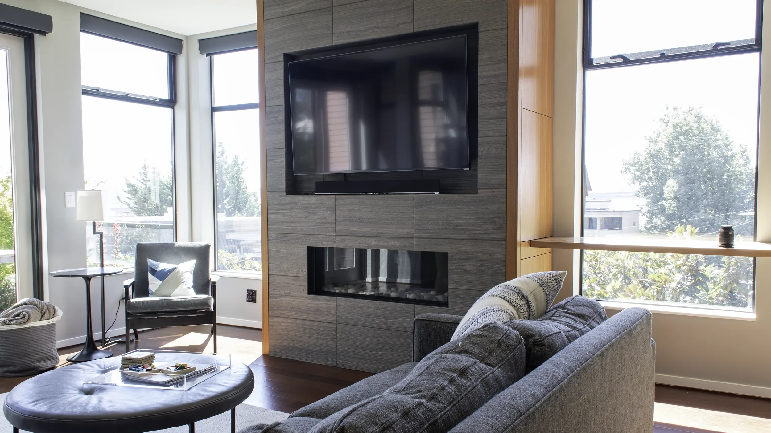 Living room with large windows, gray sofa, armchair, coffee table with magazines, wall-mounted TV above a fireplace, and a narrow wooden shelf with a small object, bright natural light.