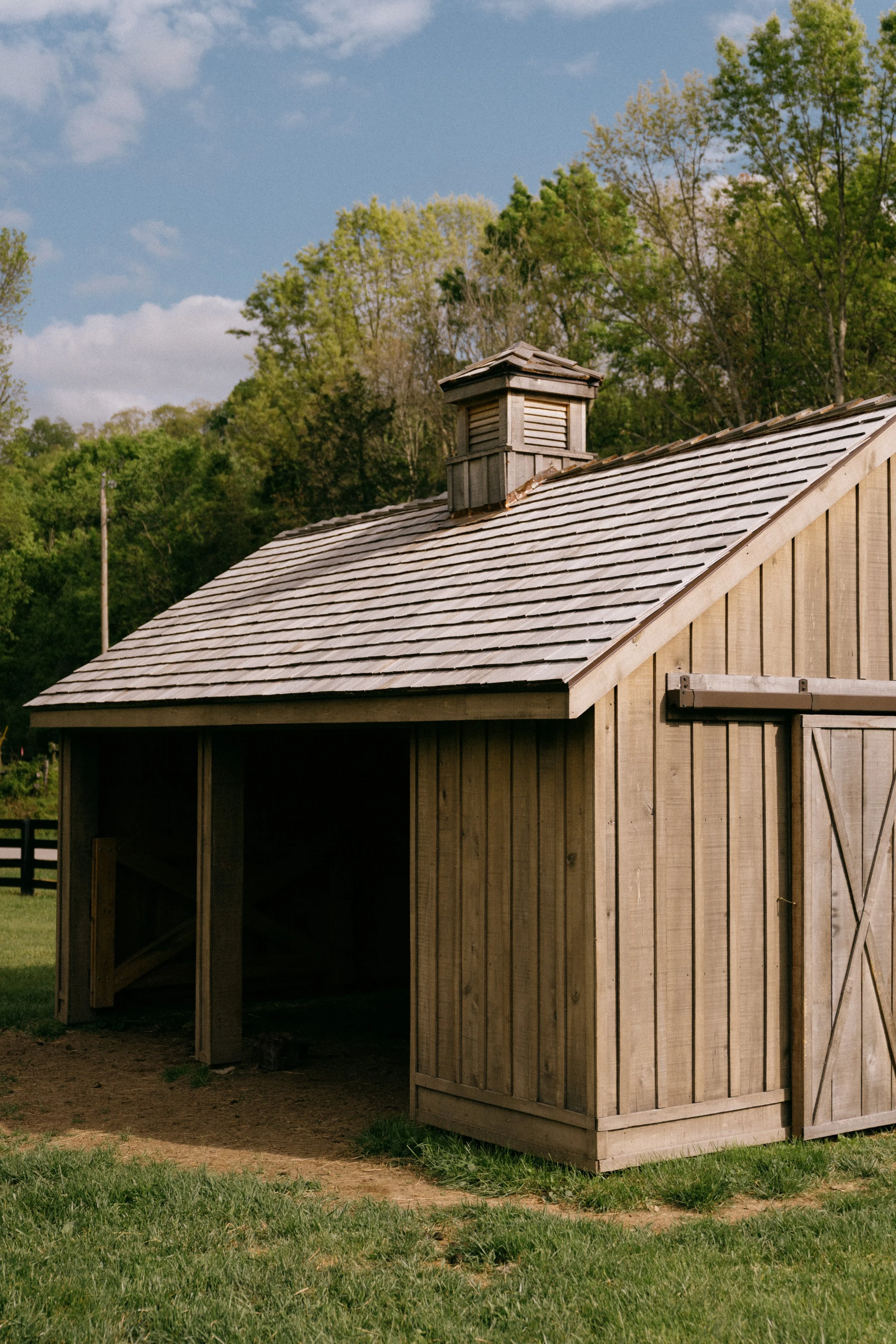 A wooden barn with a pitched roof and a small cupola on top, situated on grassy ground with trees in the background and a partly cloudy sky.