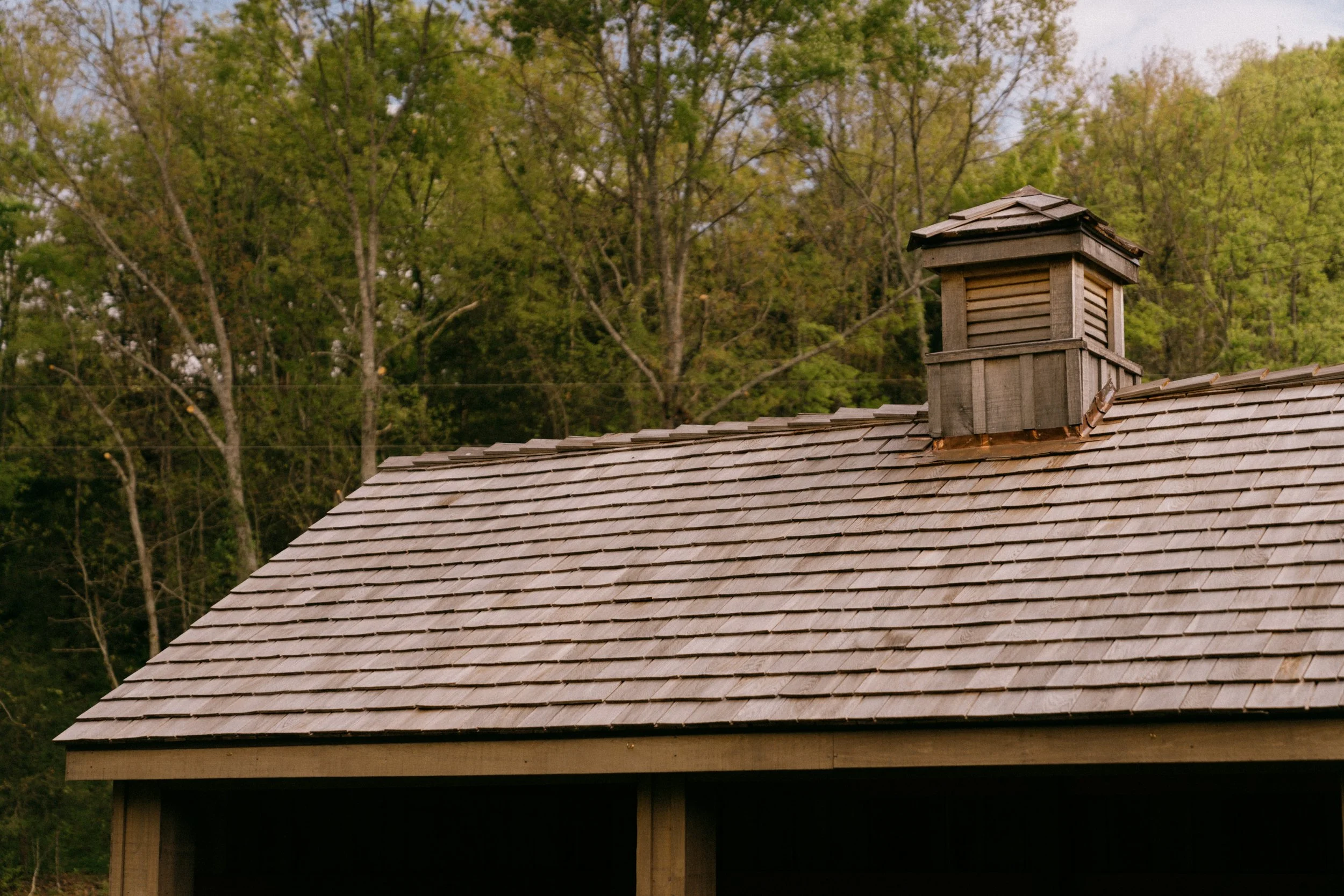 Wooden rooftop with a chimney, against a background of green trees and a cloudy sky.