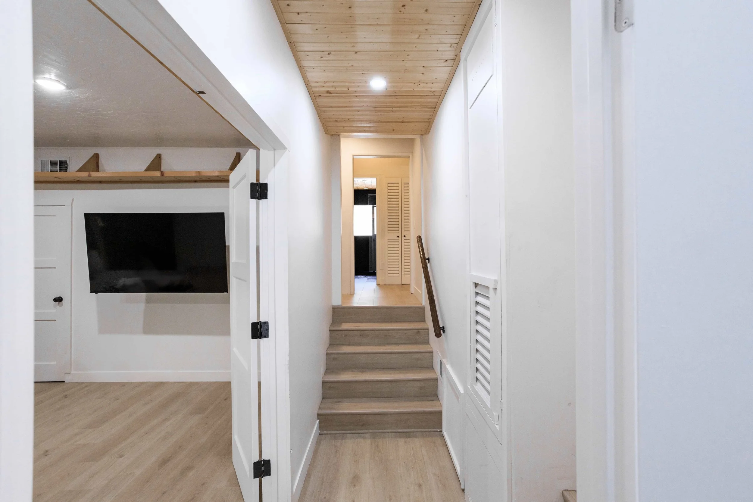 Interior hallway with light-colored wood flooring and a ceiling partially covered with wooden panels, leading to a door at the end. There are stairs with a handrail on the right side.