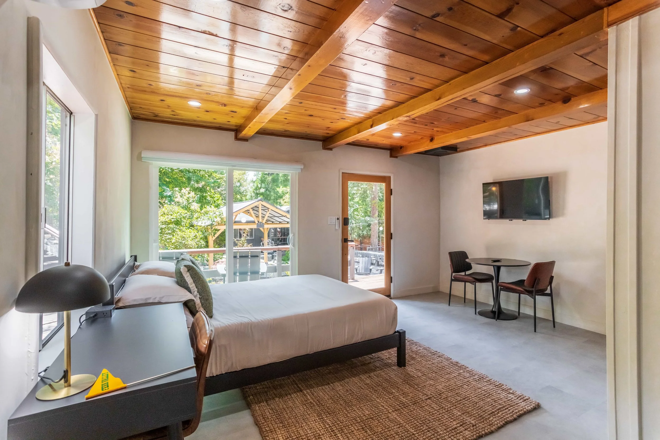 Hotel room with a bed, nightstand, lamp, sliding glass door leading to a balcony, and a wall-mounted TV. The room has wooden ceiling beams and a mix of neutral and natural decor.