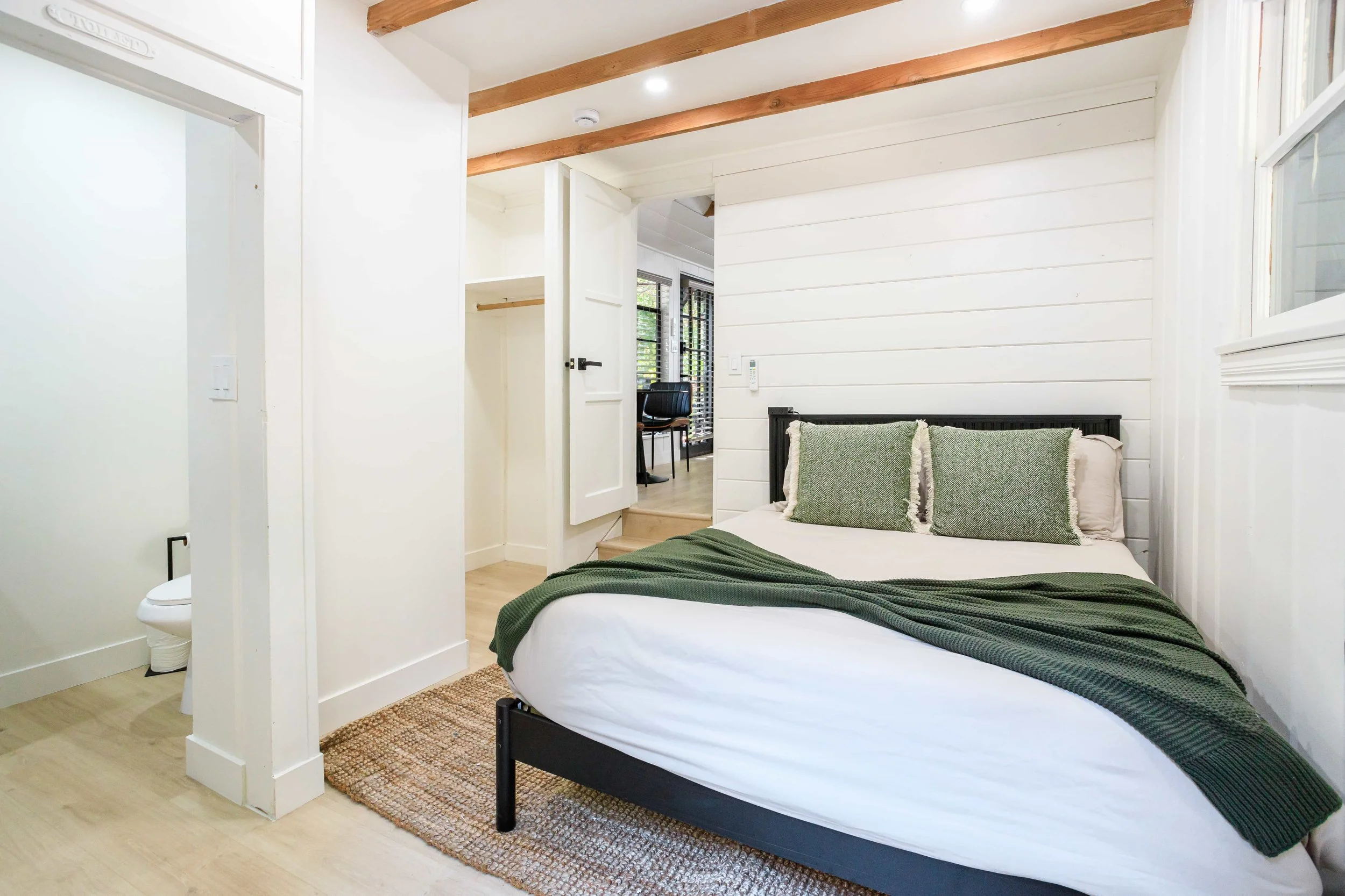 A cozy bedroom features a neatly made bed with white sheets, green pillows, and a matching green blanket. The room has white paneled walls, a woven rug, and exposed wooden beams on the ceiling. In the background, a small office nook with a desk and c