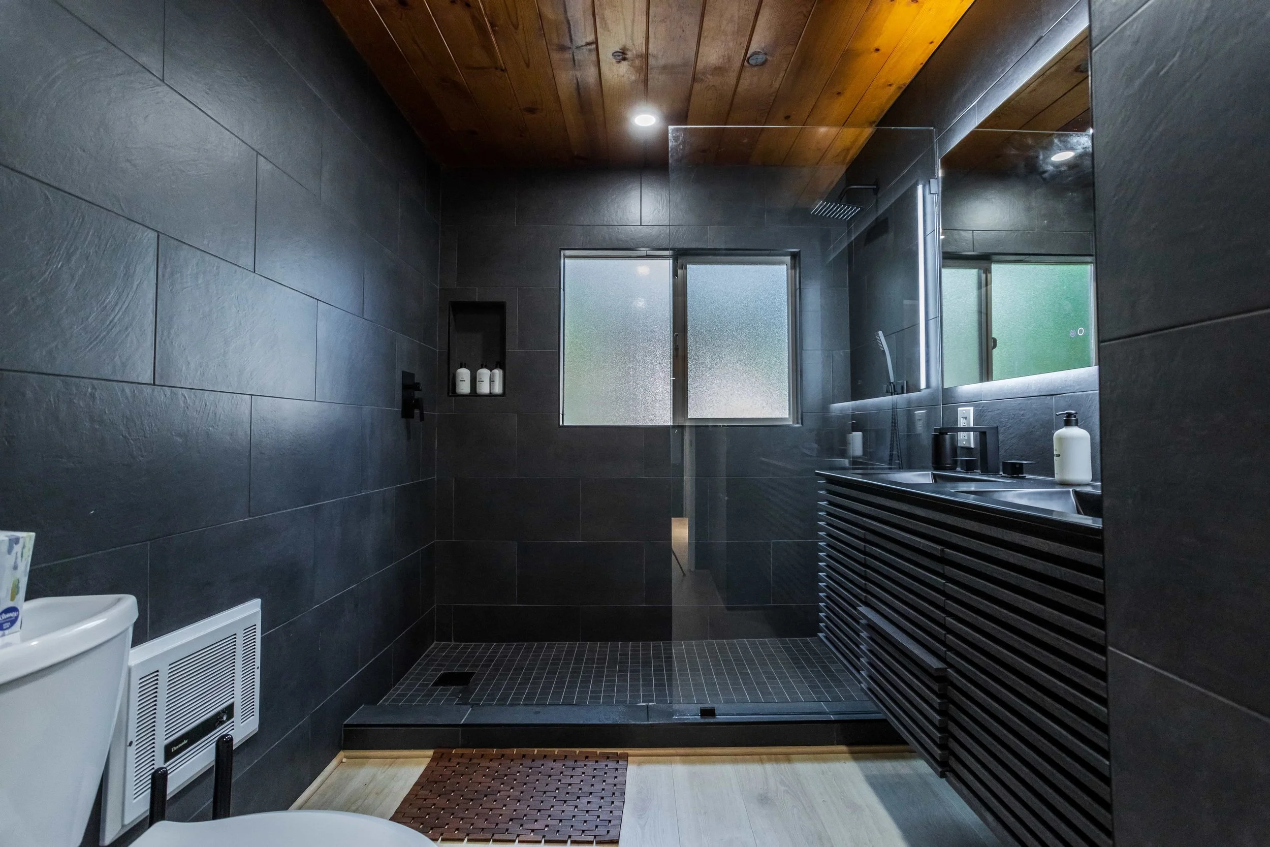 The Pinetree Hotel Idyllwild:  a - Idyllwild California, cozy cabin, mountain resort, mountain getaway Modern bathroom in cabin 1 with dark tiled walls, wooden ceiling, and walk-in shower with glass door and frosted window.