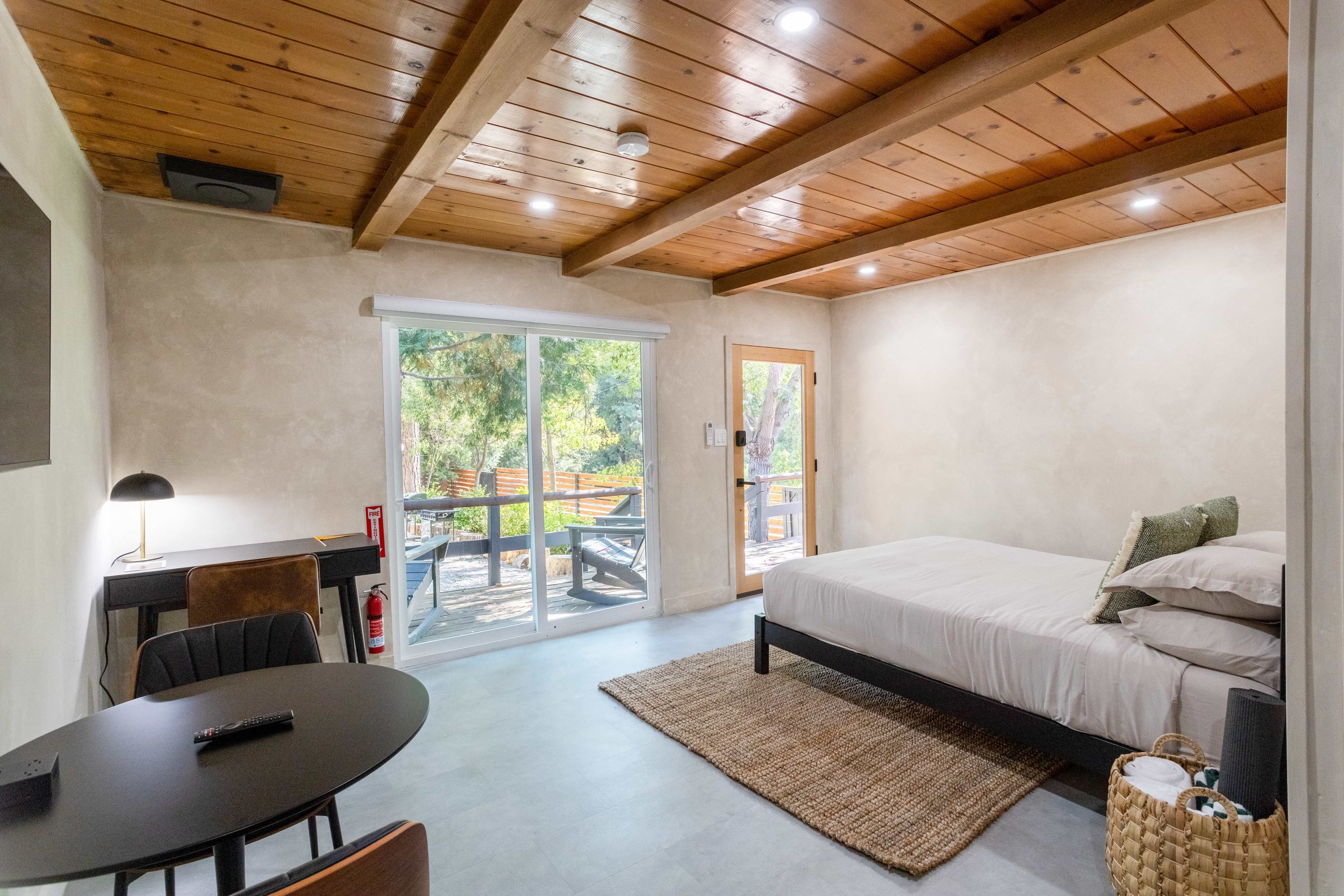 A cozy bedroom with a wooden ceiling, sliding glass door leading to an outdoor deck, single bed with white linens, a small rug, and minimal furniture.
