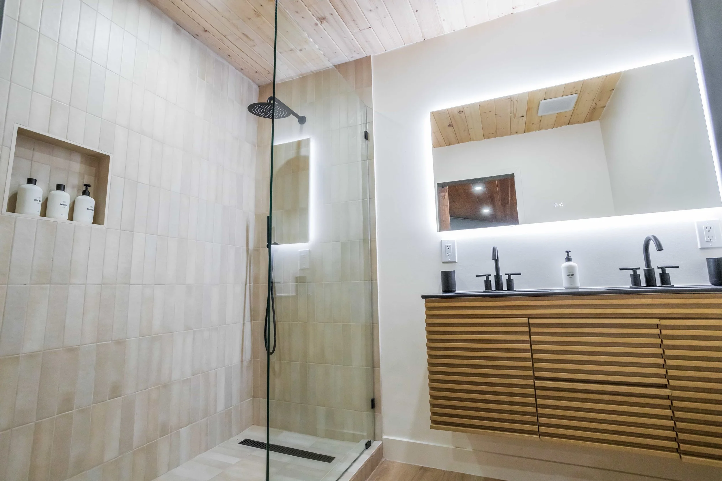 Modern bathroom with a glass-enclosed shower area and a wooden cabinet with two sinks and a large backlit mirror.