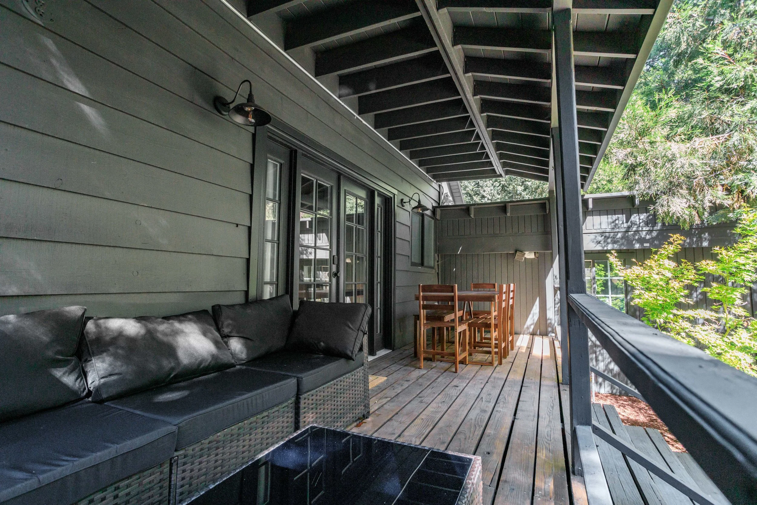 A covered wooden deck with black outdoor sofa, wooden dining table and chairs, glass door, outdoor light fixtures, and surrounded by greenery and trees.