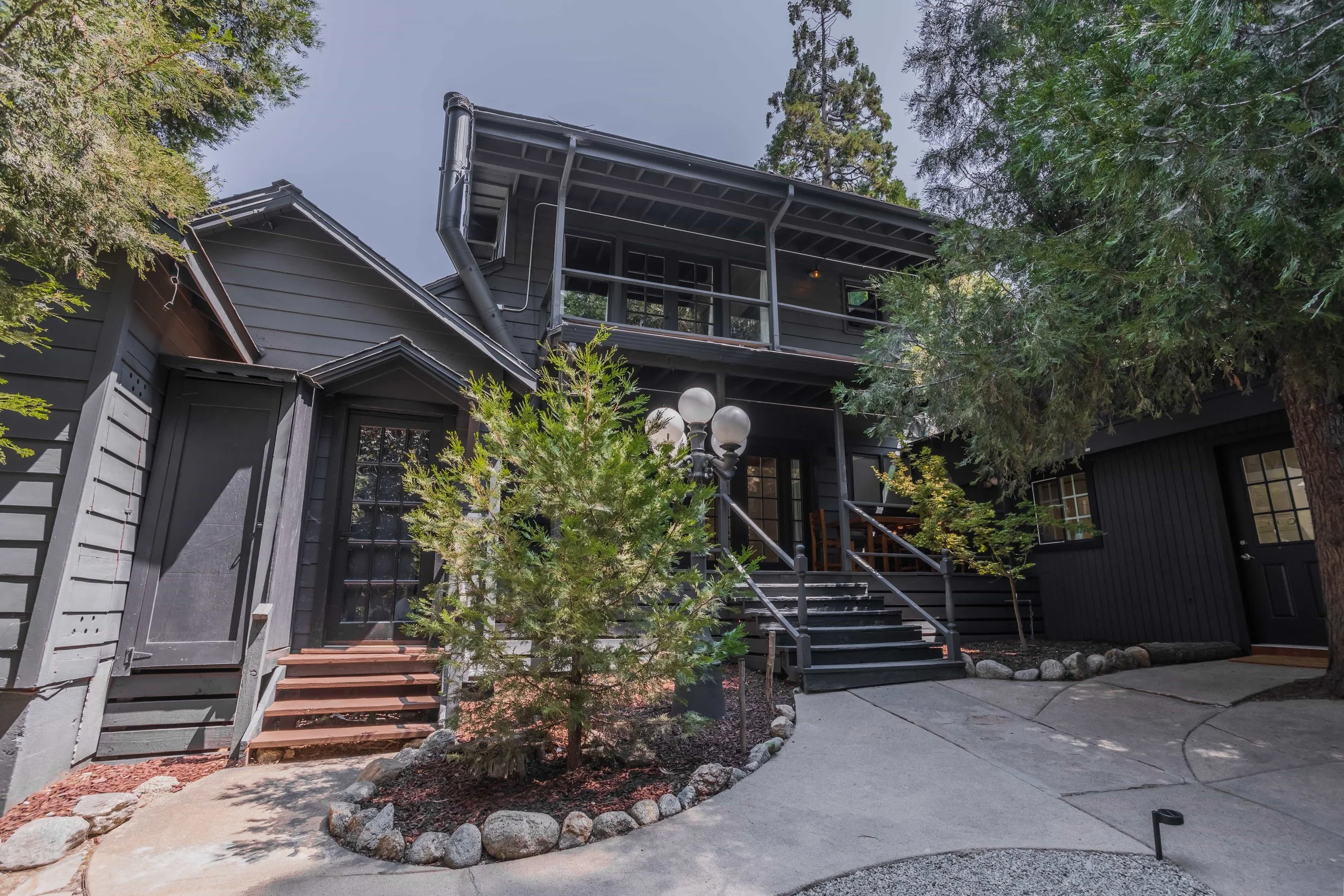 A multi-story black house with a porch, surrounded by trees and shrubs, with a concrete walkway and steps leading to the entrance.