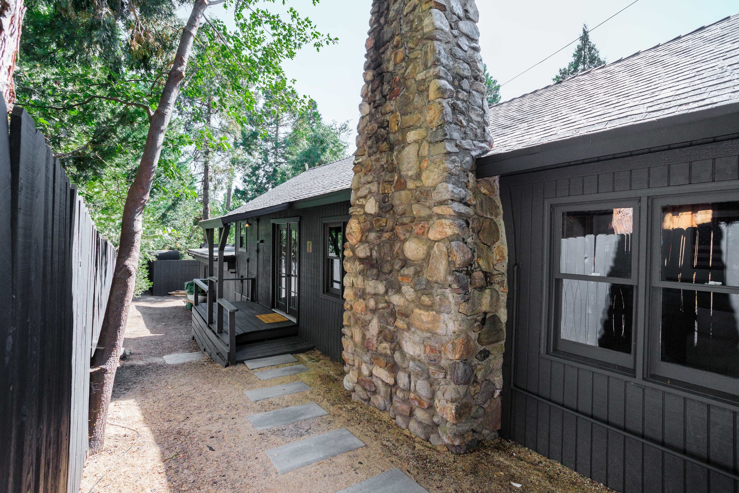 Side view of a black wooden house with a stone chimney, set in a wooded area. A small porch with steps and a welcome mat is visible. The yard has a gravel path with stepping stones, and a tall black fence borders the property.
