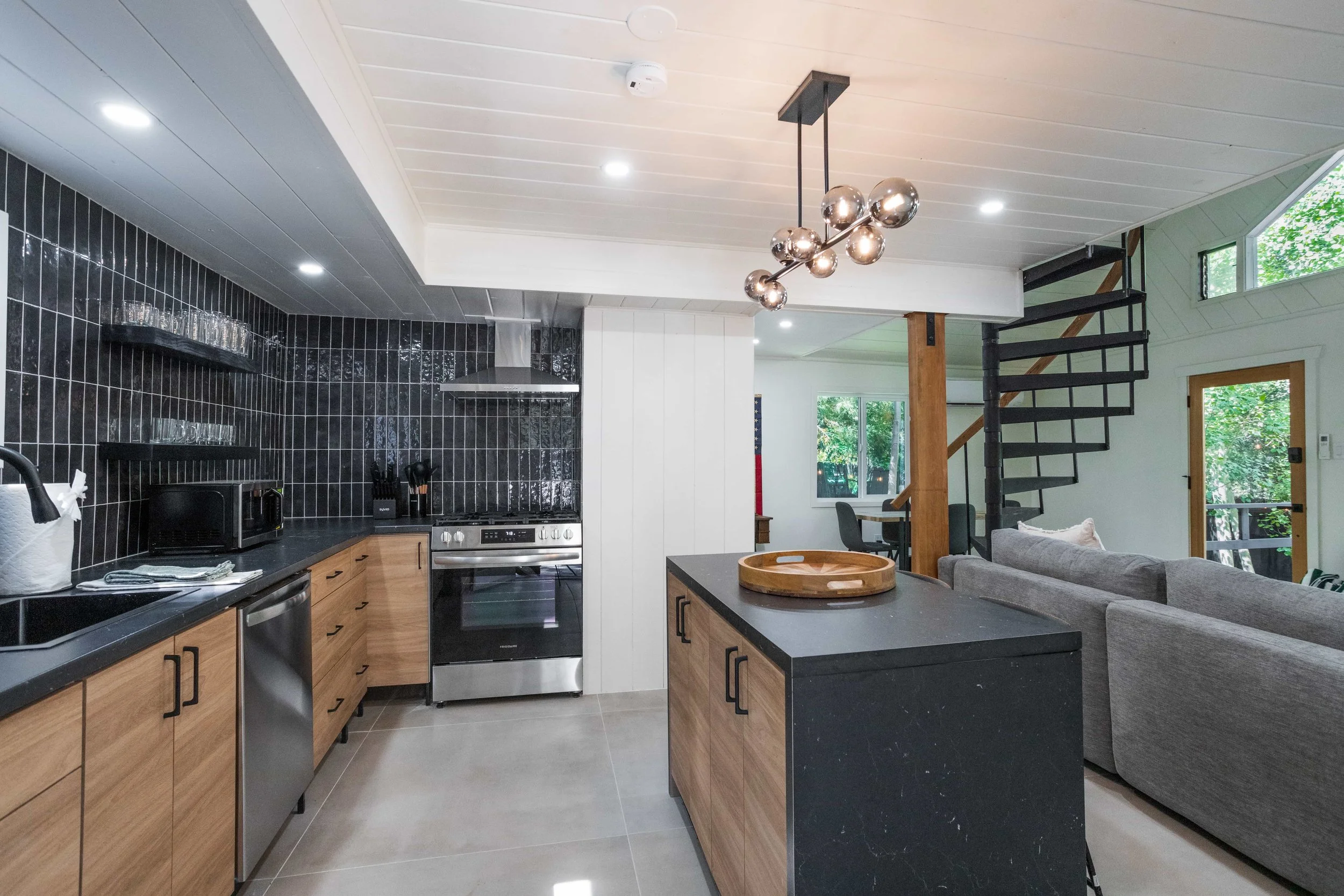Open-concept kitchen and living room with black tiled backsplash, wooden cabinets, modern appliances, a central island with a tray, and a gray sectional sofa. Spiral staircase with black steps leads to upper floor, large windows bringing in natural l