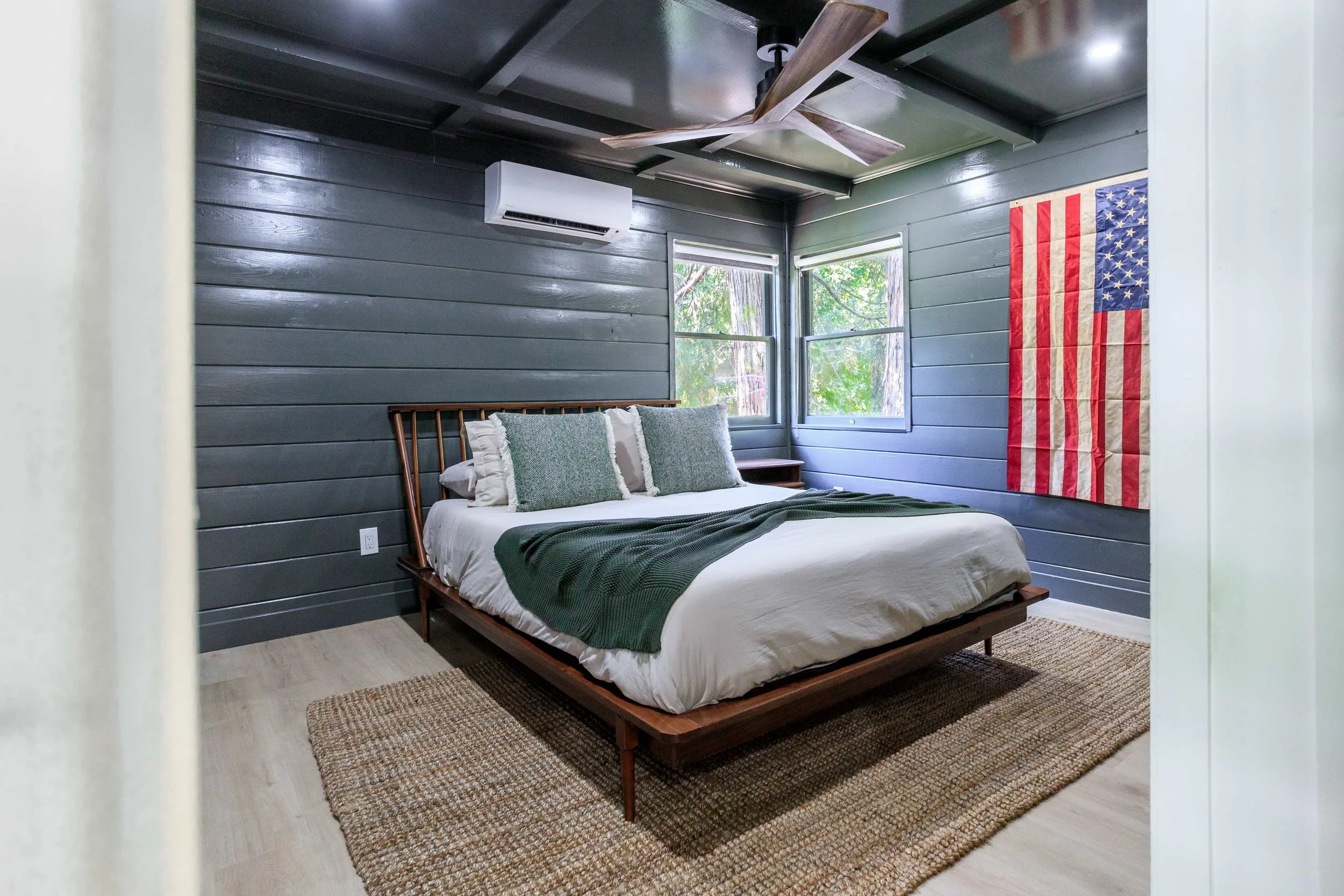 A bedroom with dark gray wood-paneled walls, a wooden bed frame with white bedding and green accent pillows, a beige rug, a ceiling fan, an air conditioning unit, and a U.S. flag hanging on the wall.