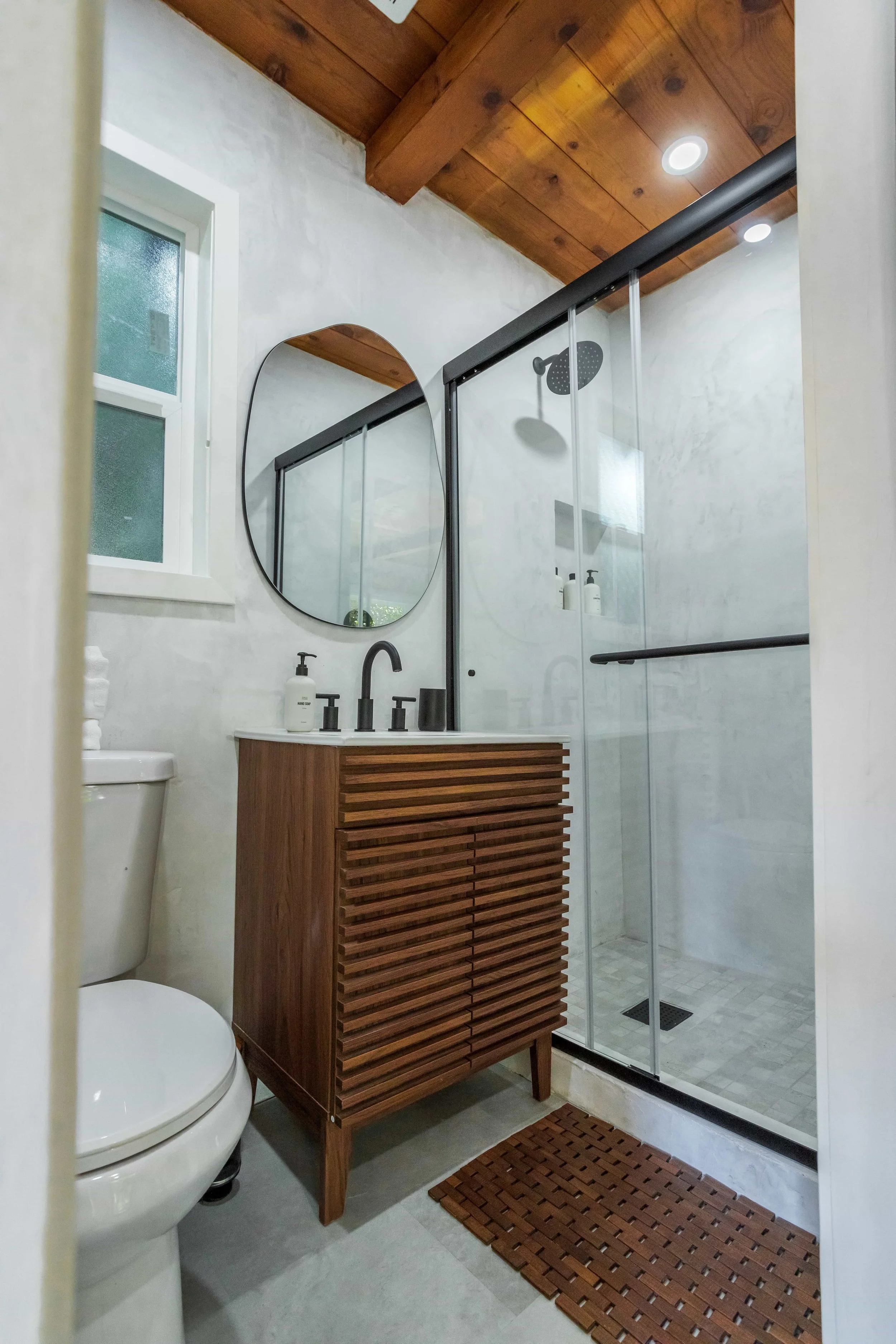 A modern bathroom featuring a white toilet, a wooden vanity with black fixtures, a round mirror, a frosted window, and a glass shower with a black frame and wooden ceiling.