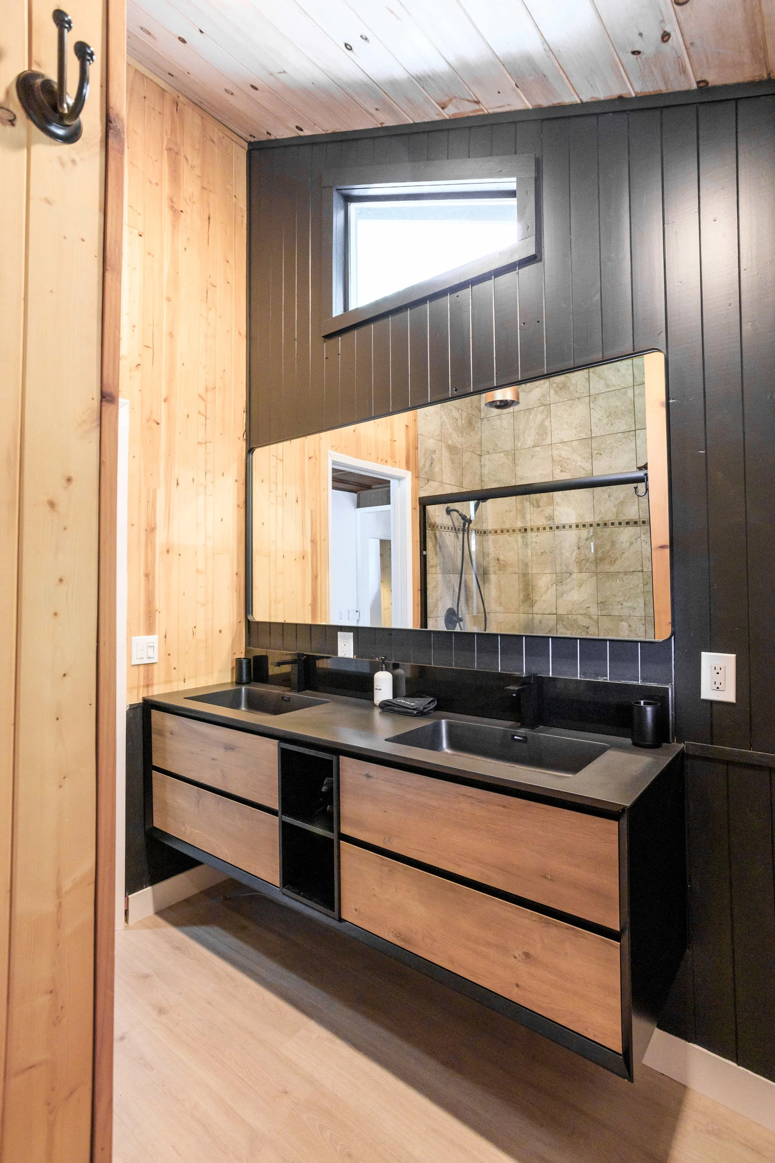 Modern bathroom vanity with dual sinks, black countertops, and wooden drawers, large mirror, and a window in a wood-paneled room.