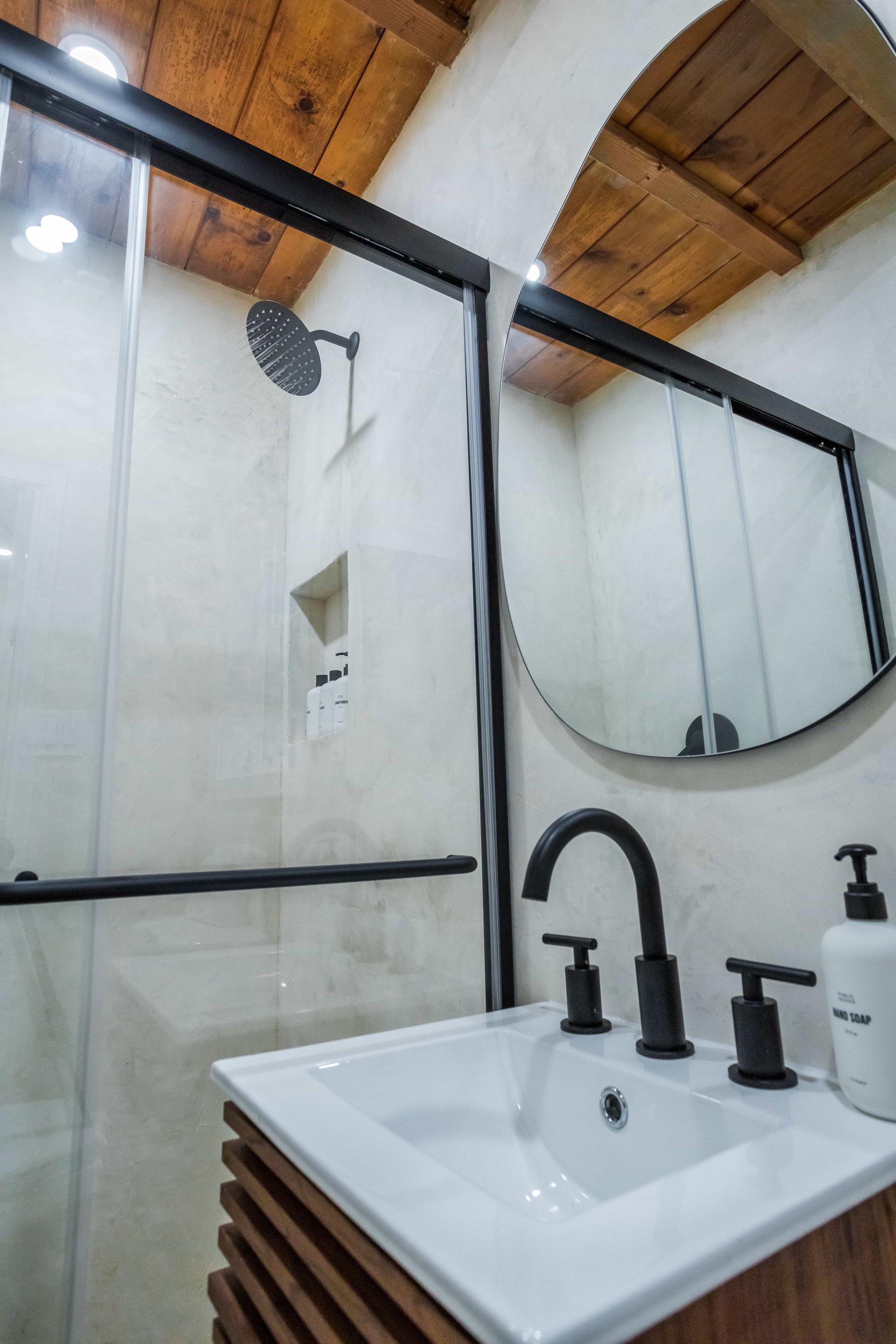 Modern bathroom with a black shower head, glass shower enclosure, round mirror, black faucet, and soap dispenser.