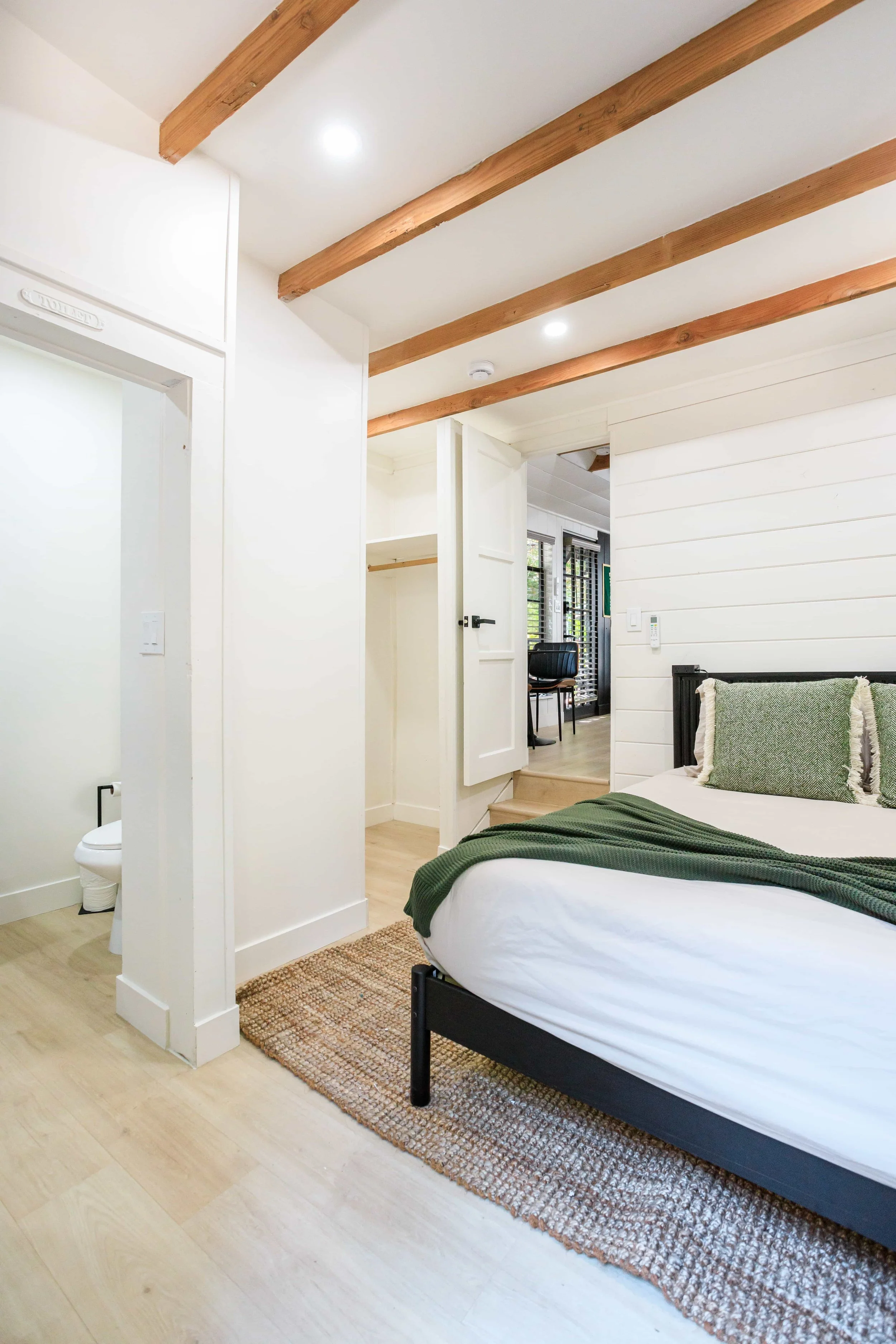 Bedroom with a bed dressed in white linens and a green blanket, an open closet door, and a partial view of a bathroom with a toilet.