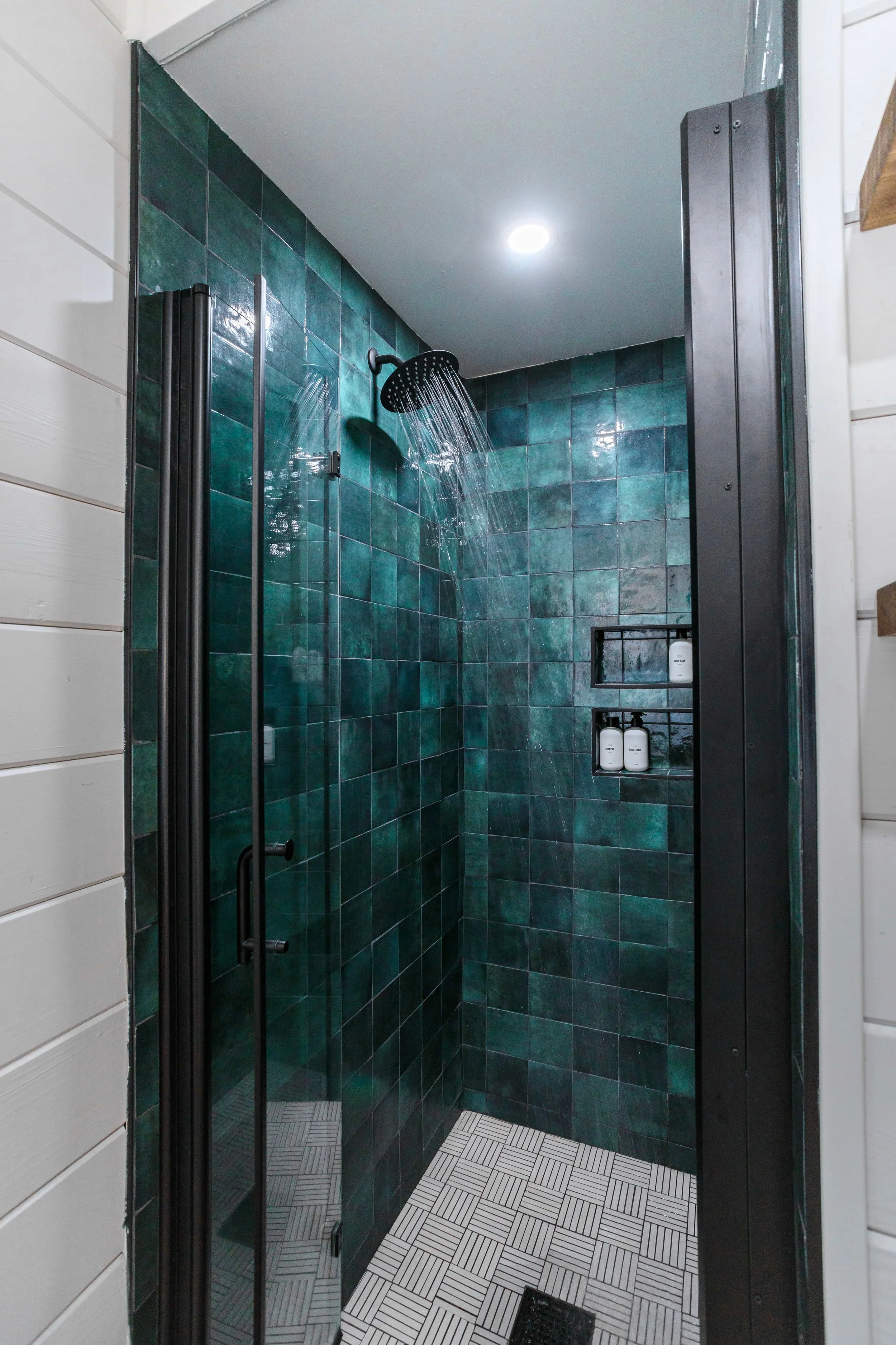 A modern shower with dark green tiled walls, a black showerhead, and built-in shelves holding bottles, with a white ceiling and a recessed light.