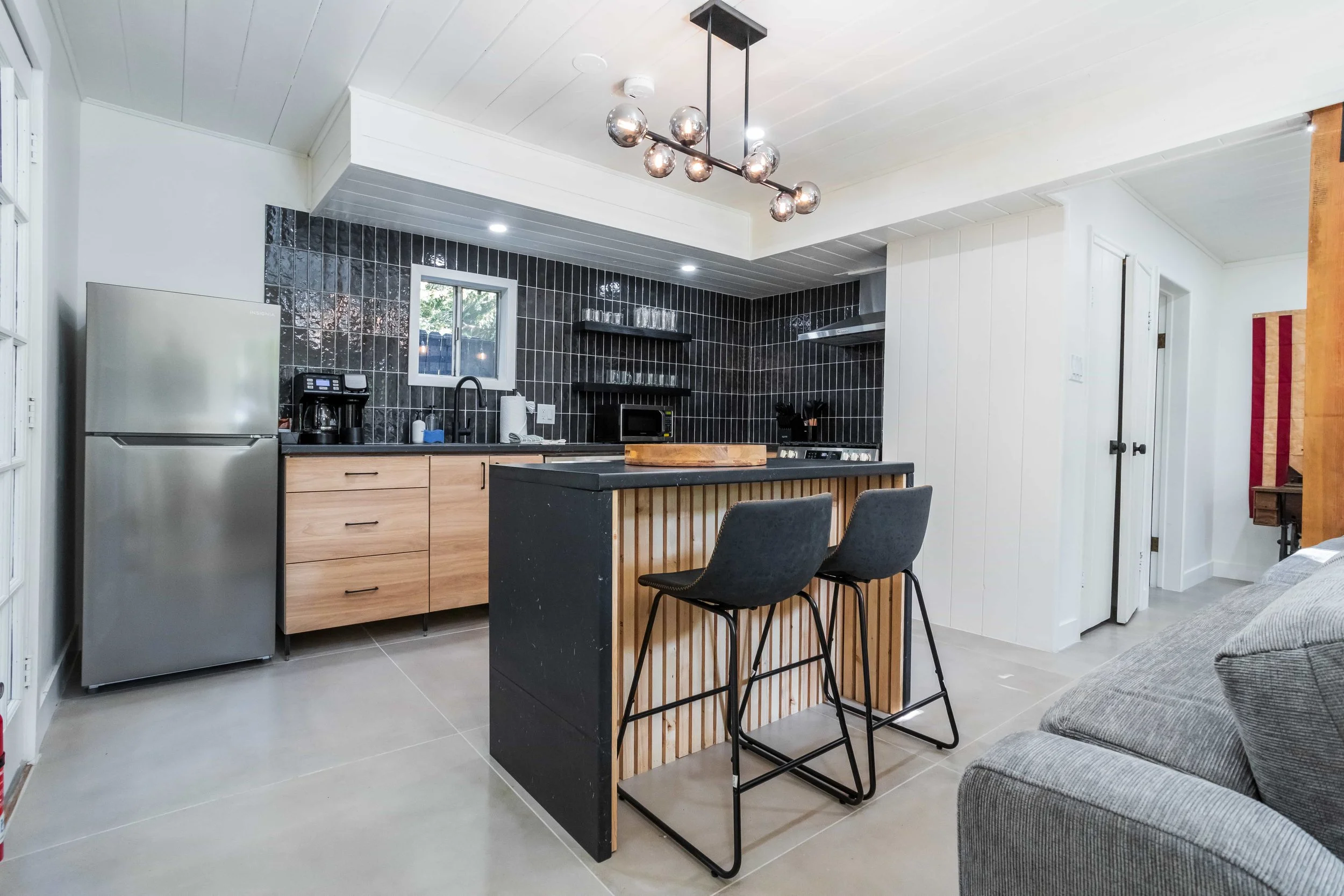 Modern kitchen with black tile backsplash, light wood cabinets, stainless steel refrigerator, black counter with stools, and a black island with wood slat accents. Contains a coffee maker, microwave, sink, and hanging light fixture.