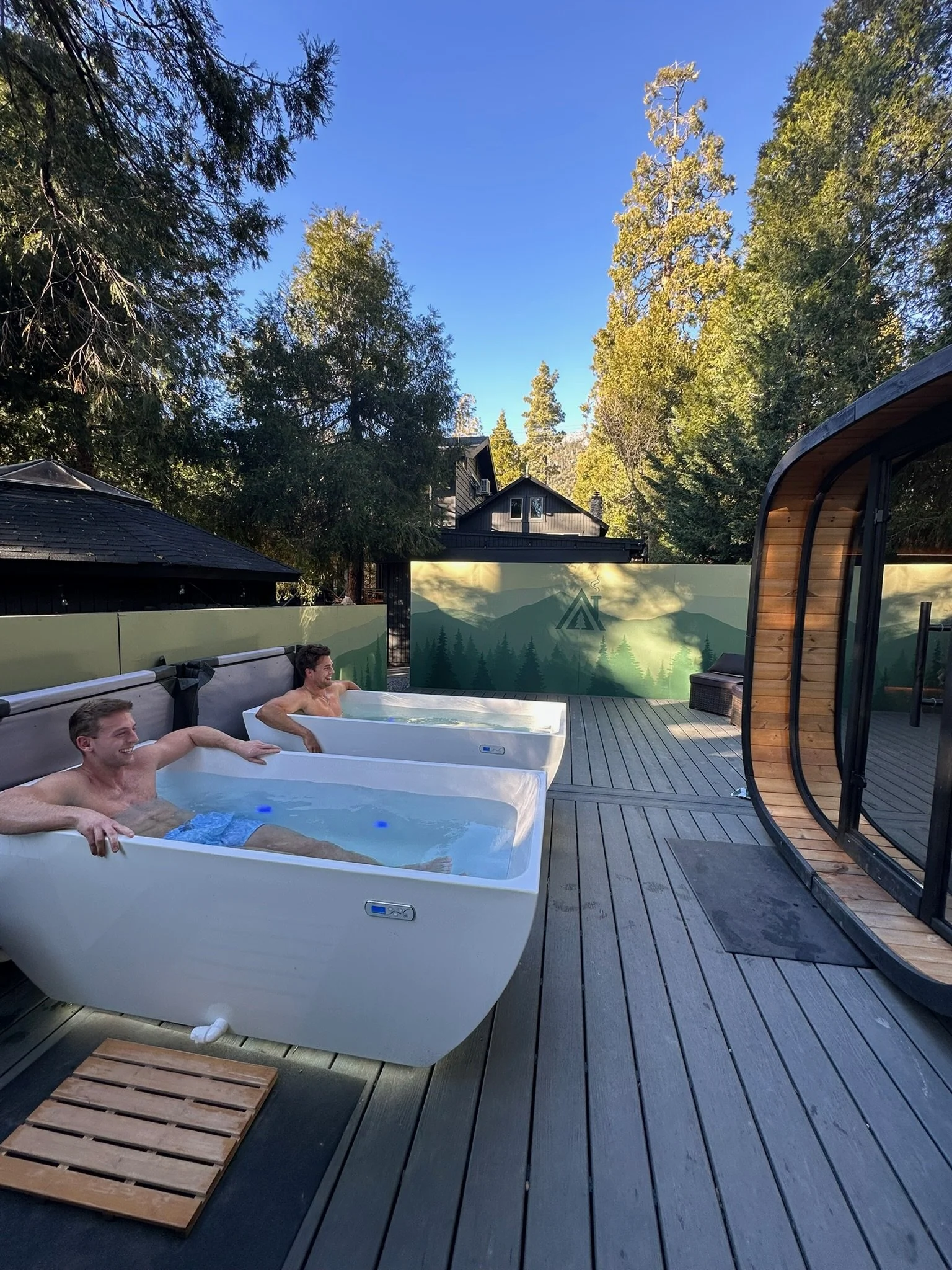 Two men relaxing in hot tubs on a wooden deck outdoors with trees and a house in the background during daytime.