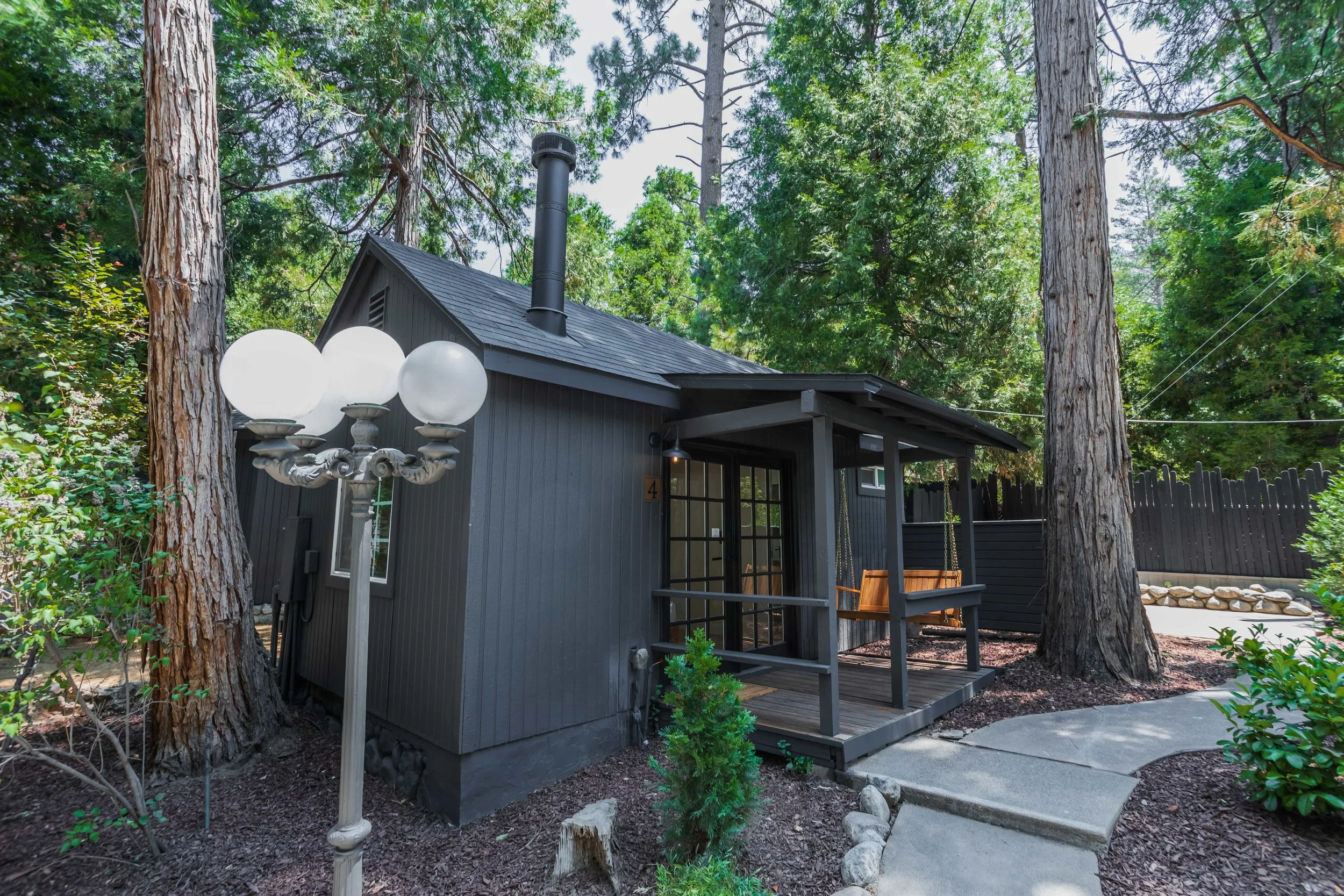 Small black wooden house with front porch, swing, large trees, and vintage street lamp in a lush green garden.