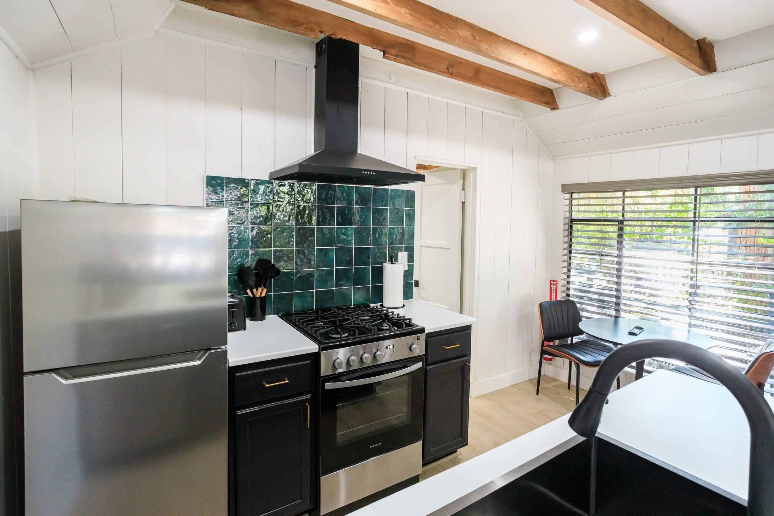 Kitchen area with a stainless steel refrigerator, black cabinets, green tiled backsplash, gas stove, paper towel holder, window with blinds, and a small table with chairs.