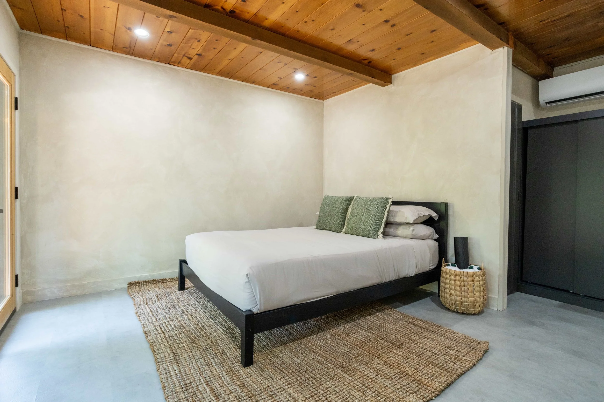 A minimalist bedroom with a black metal bed frame, white bedding, and two green pillows. The room has a wooden ceiling, beige walls, a woven rug, and a basket beside the bed. An air conditioner is mounted on the wall.