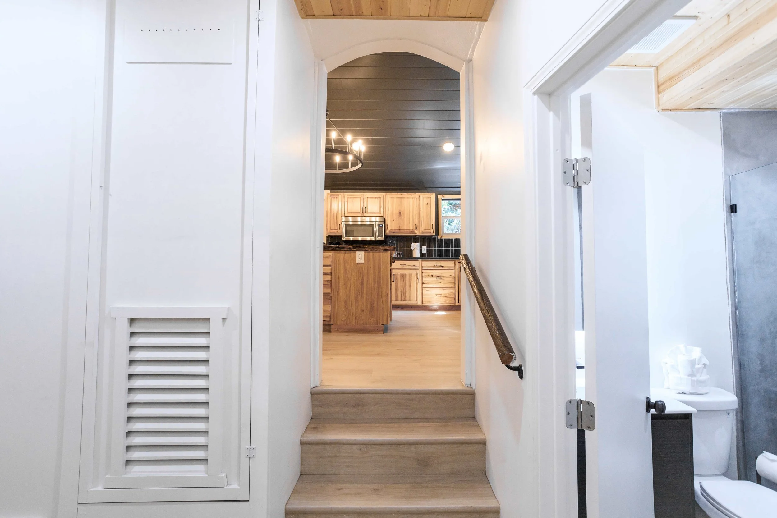View of a staircase leading up to a kitchen with wooden cabinets, black backsplash, and a stainless steel microwave, seen through an arched doorway from a hallway with a handrail and a bathroom with a toilet on the right.