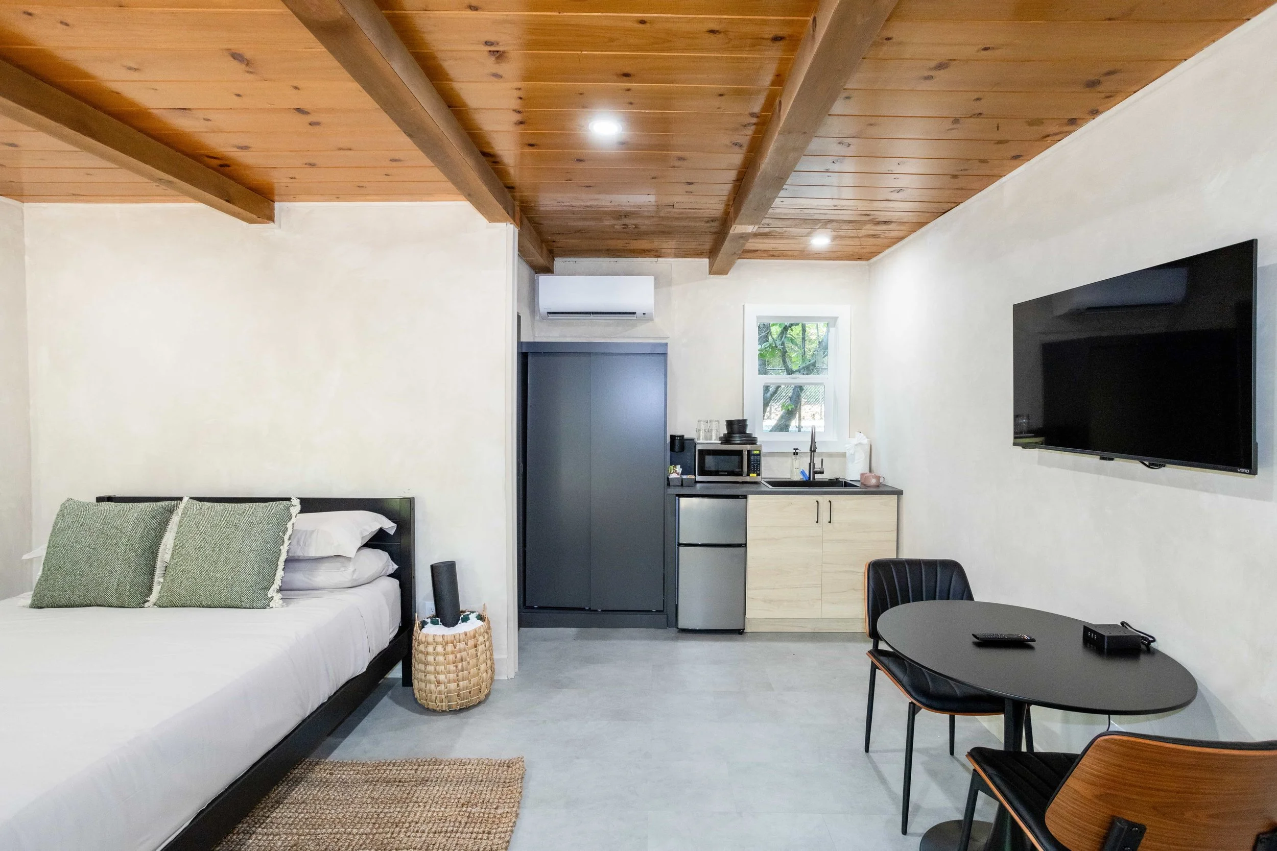Interior of a minimalist studio apartment with a bed, small kitchenette, wall-mounted television, and a dining table with chairs.