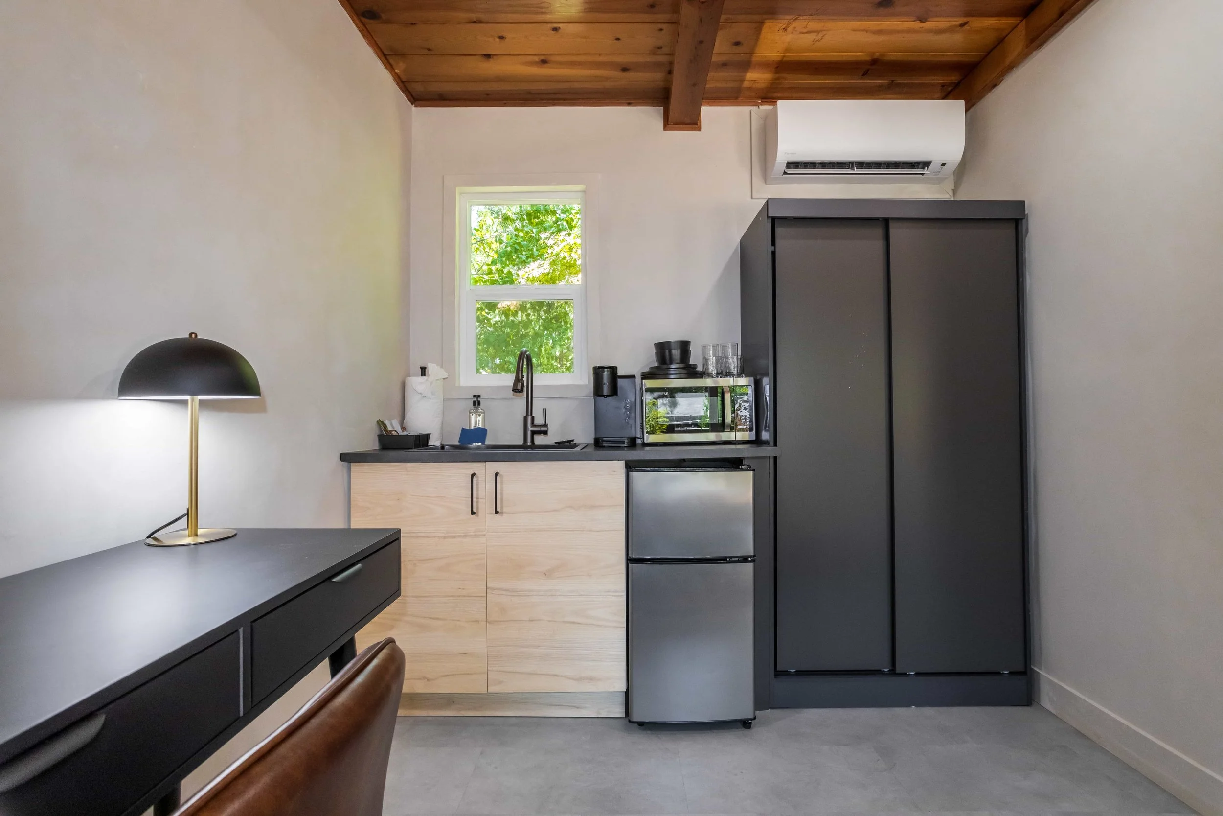 Small kitchen with wooden ceiling, beige walls, black desk with lamp, compact fridge, black storage cabinet, microwave, coffee maker, and a window with green trees outside.