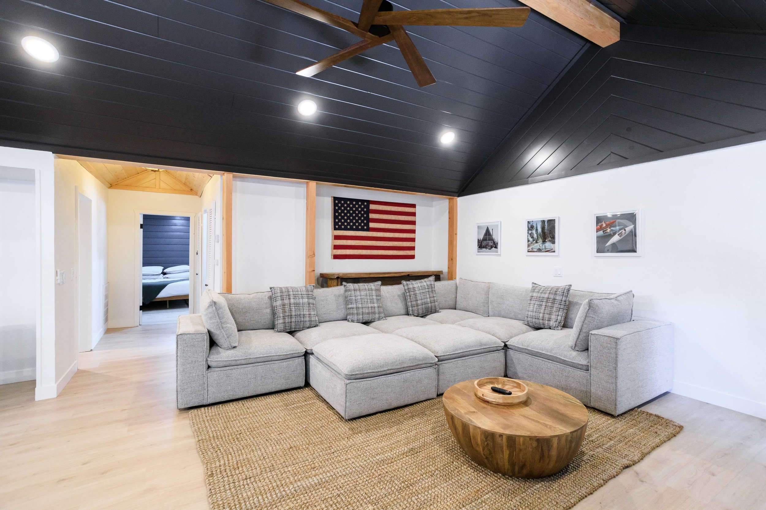 Living room with a large gray sectional sofa, patterned pillows, a round wooden coffee table, framed pictures on the wall, an American flag hanging on the wall, and a bedroom visible through an open doorway. The ceiling is dark with recessed lighting