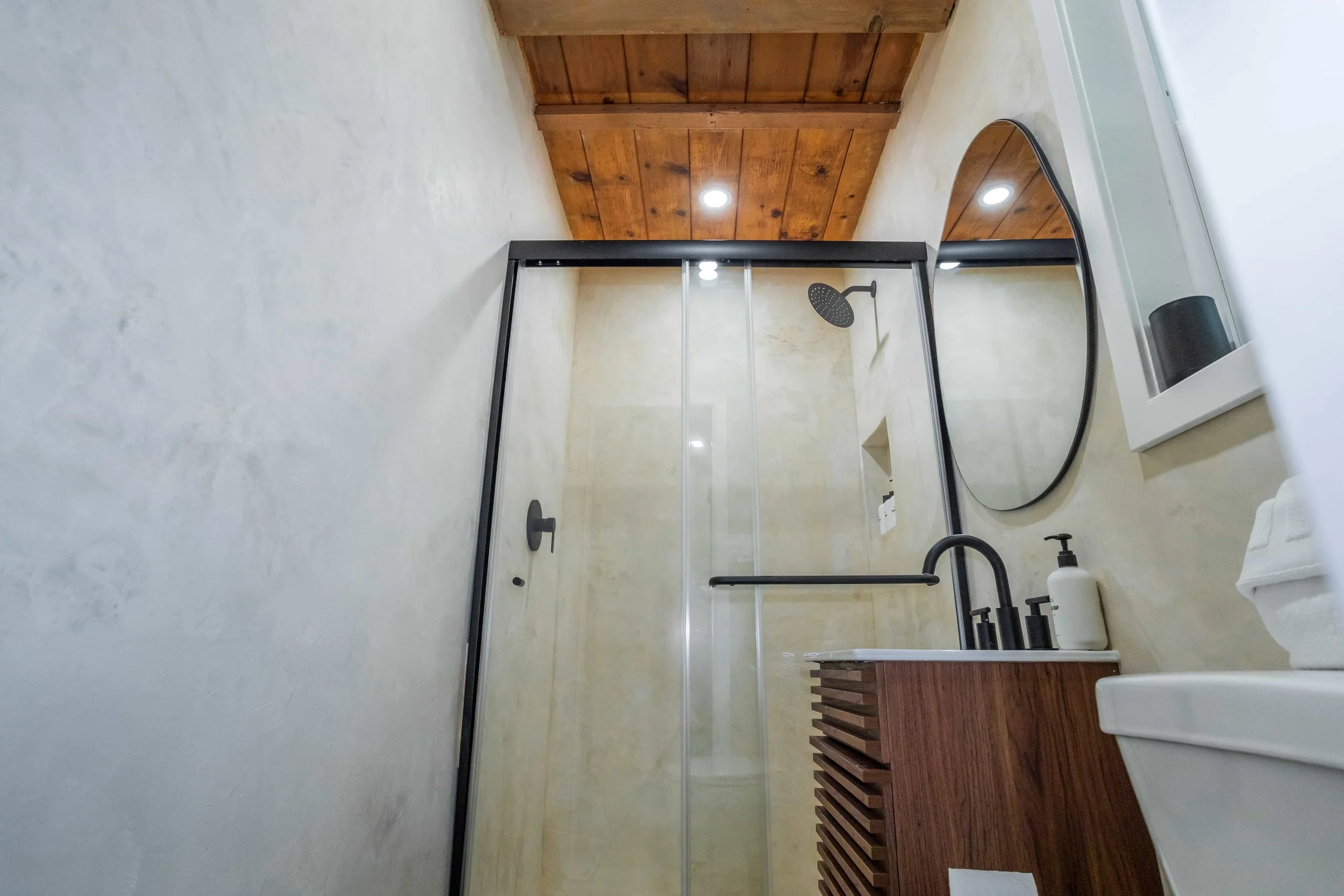 View of a modern bathroom with a walk-in shower featuring a glass door and black fixtures, a wooden ceiling, a rounded mirror, a wooden vanity with a white countertop and black faucet, a wall-mounted liquid soap dispenser, and a white toilet.