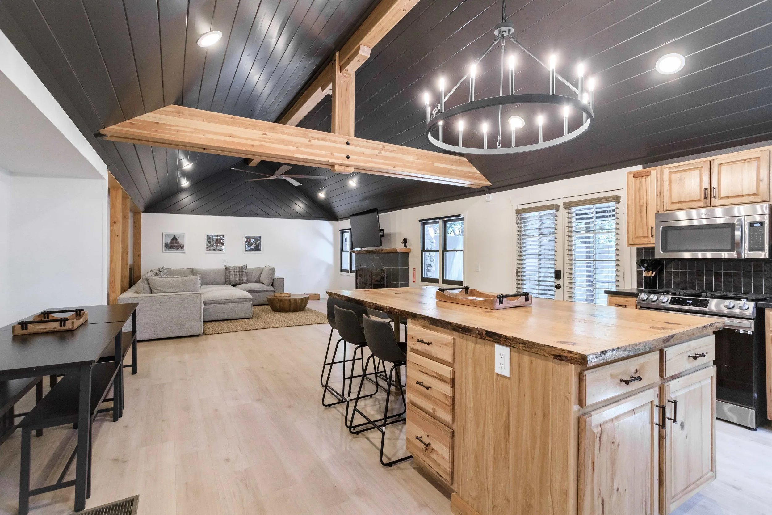 Open-concept living room and kitchen with wooden accents, black ceiling, modern lighting, gray sectional sofa, and large windows.