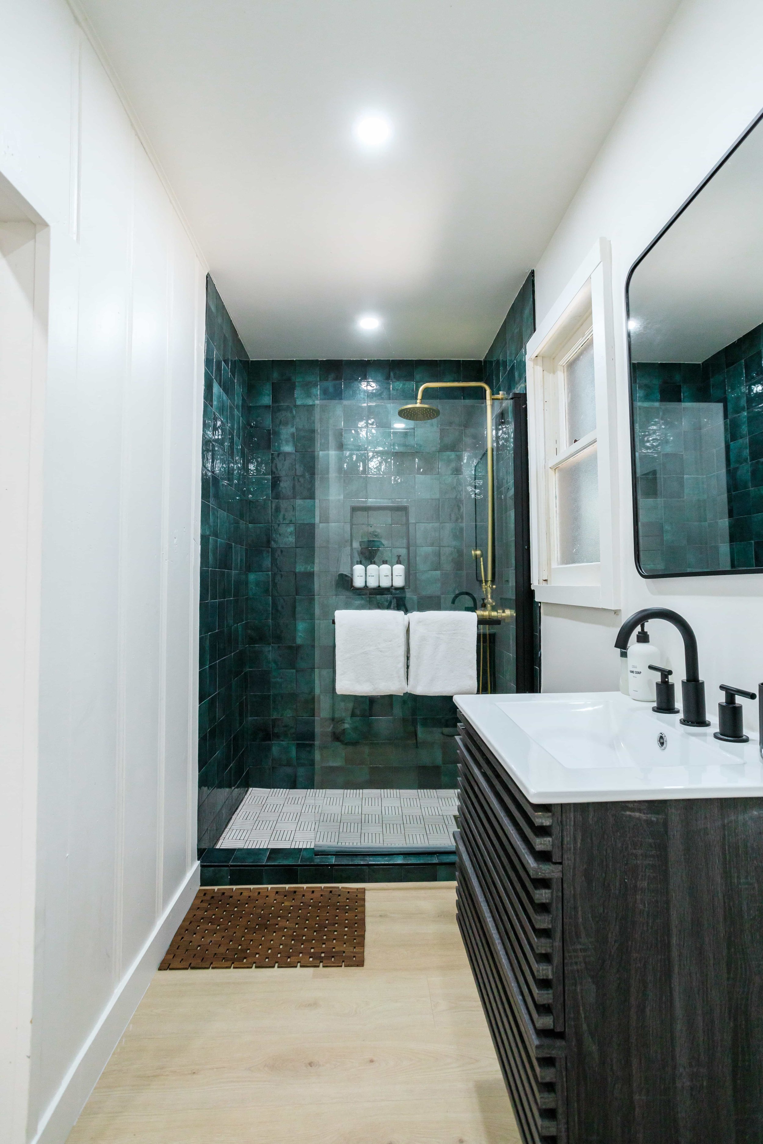Modern bathroom with a walk-in shower featuring dark green tiles, a white sink with black fixtures, and a small window.