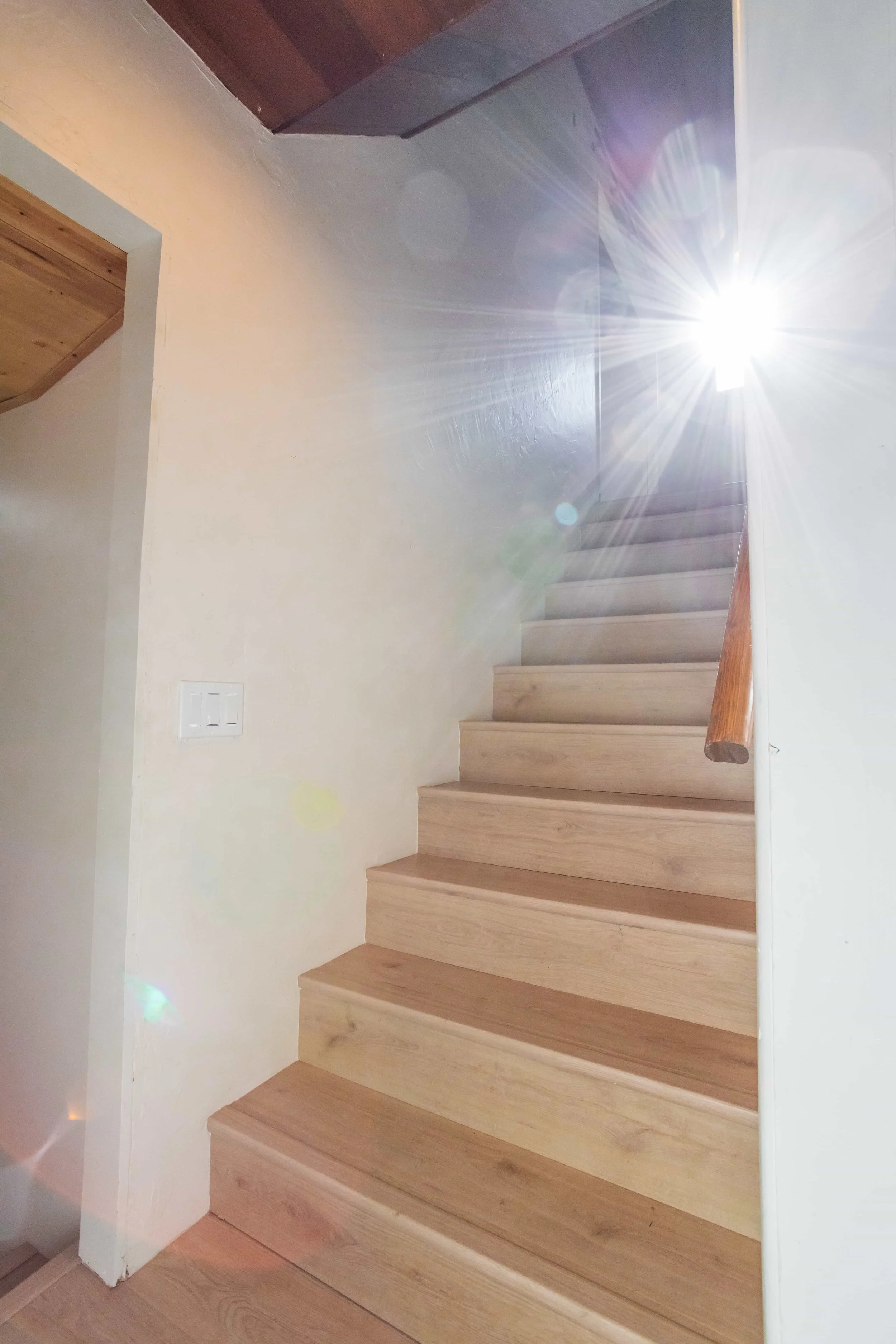 Wooden staircase with a simple wooden handrail, sunlight streaming in from a window above, sun flare and lens flares visible in the photo.