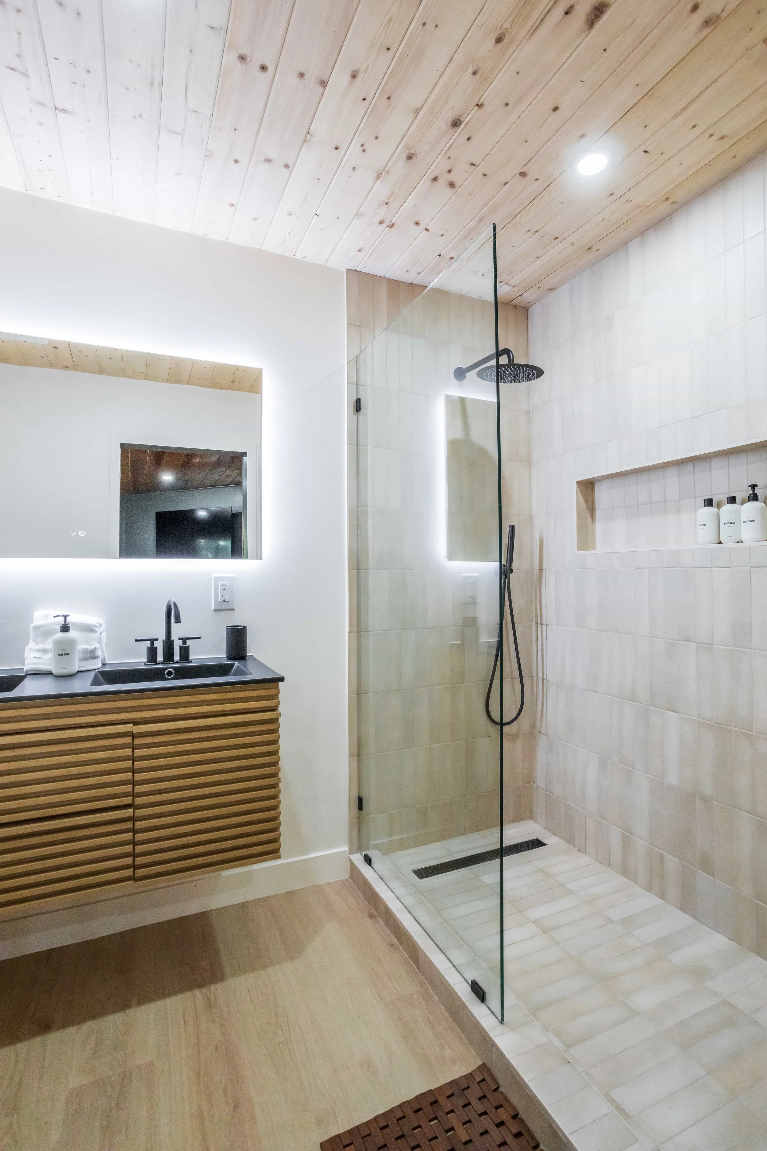 A modern bathroom with a wooden ceiling, a black sink vanity with a large mirror, a walk-in shower with beige tile walls, a black rainfall showerhead, a handheld shower, and a built-in wall niche with bottles. There is a glass partition separating th