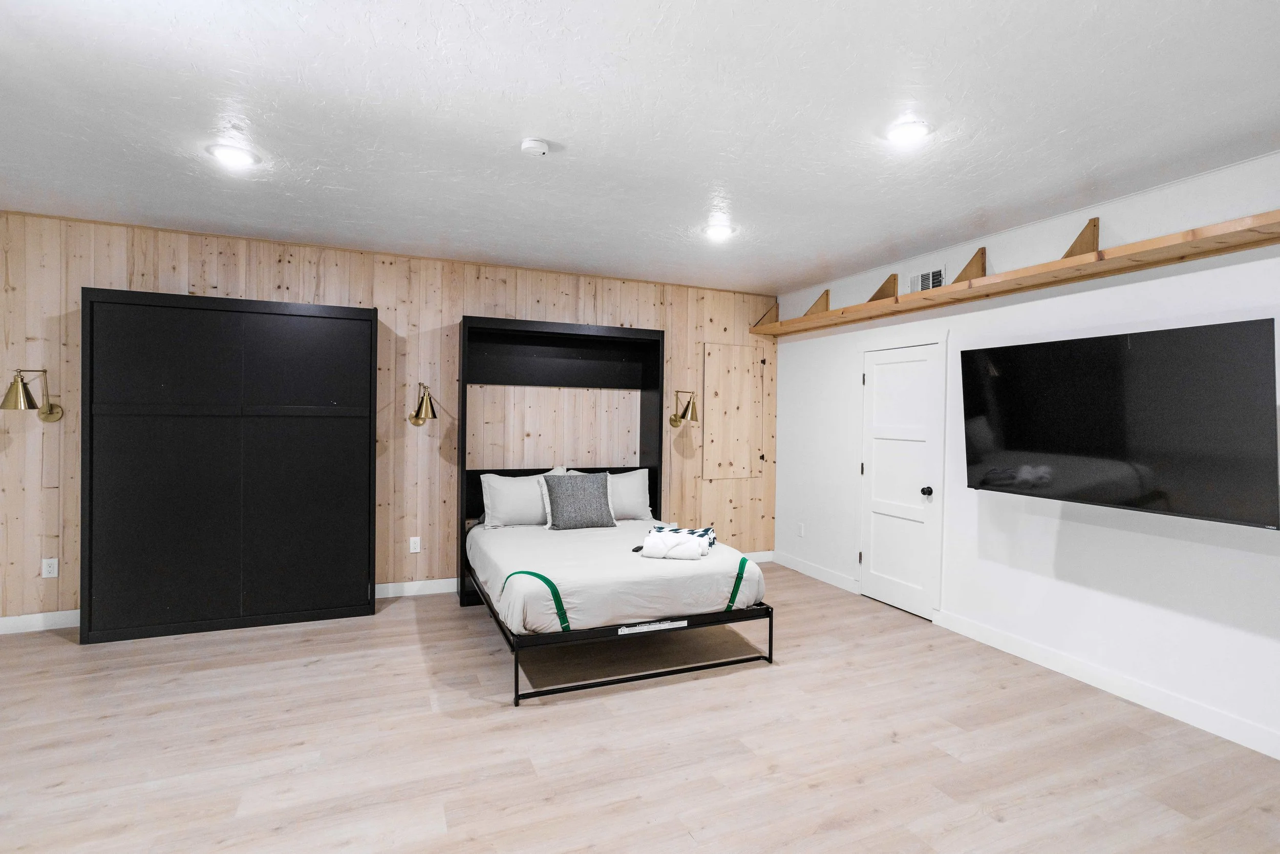 A minimalist bedroom with a wooden accent wall, black headboard, white bedding, a flat-screen TV on the white wall, and a single bed with a gray pillow and folded towel on it.