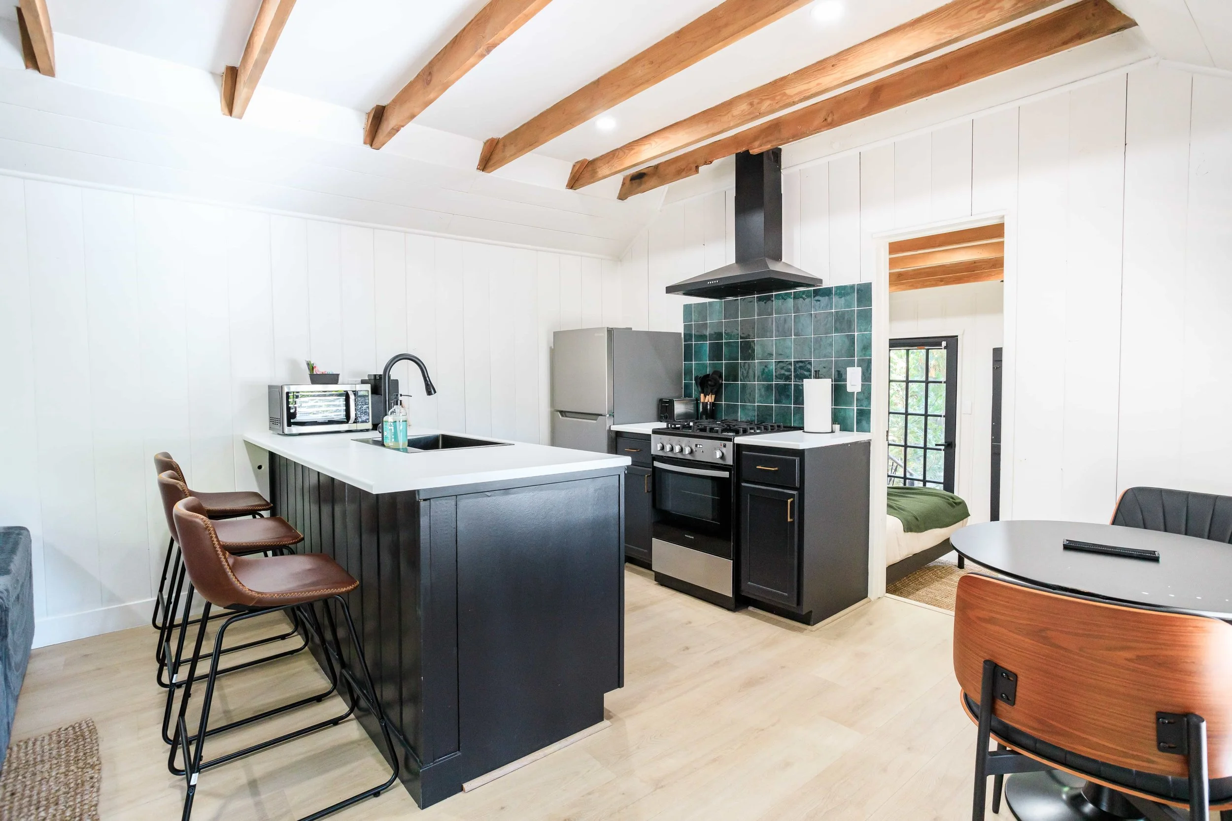 Modern kitchen with black cabinets, stainless steel appliances, green tiled backsplash, and white walls. There is a kitchen island with brown bar stools, a round dining table, and a bedroom visible through an open doorway.