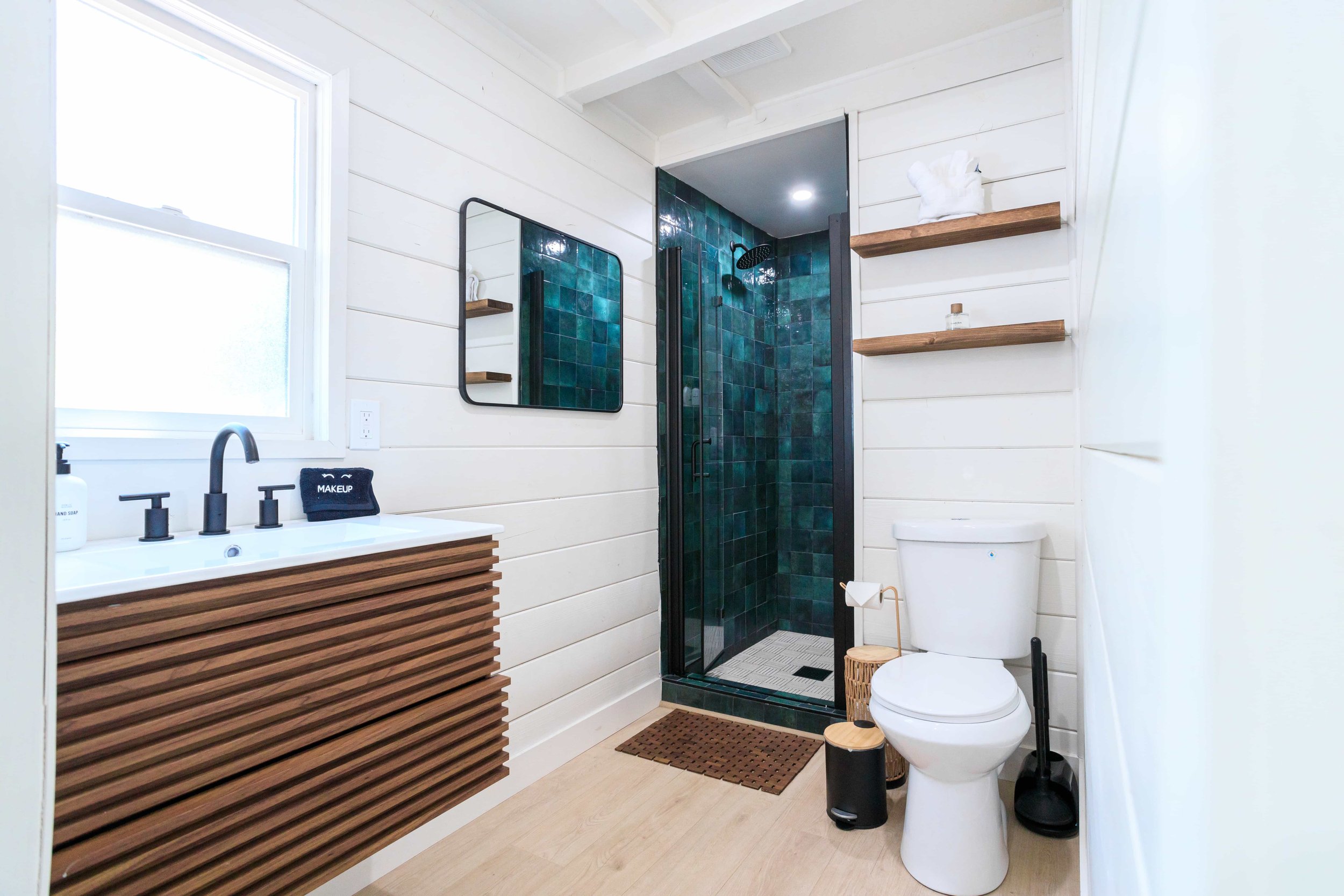 Modern bathroom with white wooden walls, a shower with dark green tiles, a wooden vanity with black fixtures, and floating wooden shelves. Includes a toilet, mirror, and window.