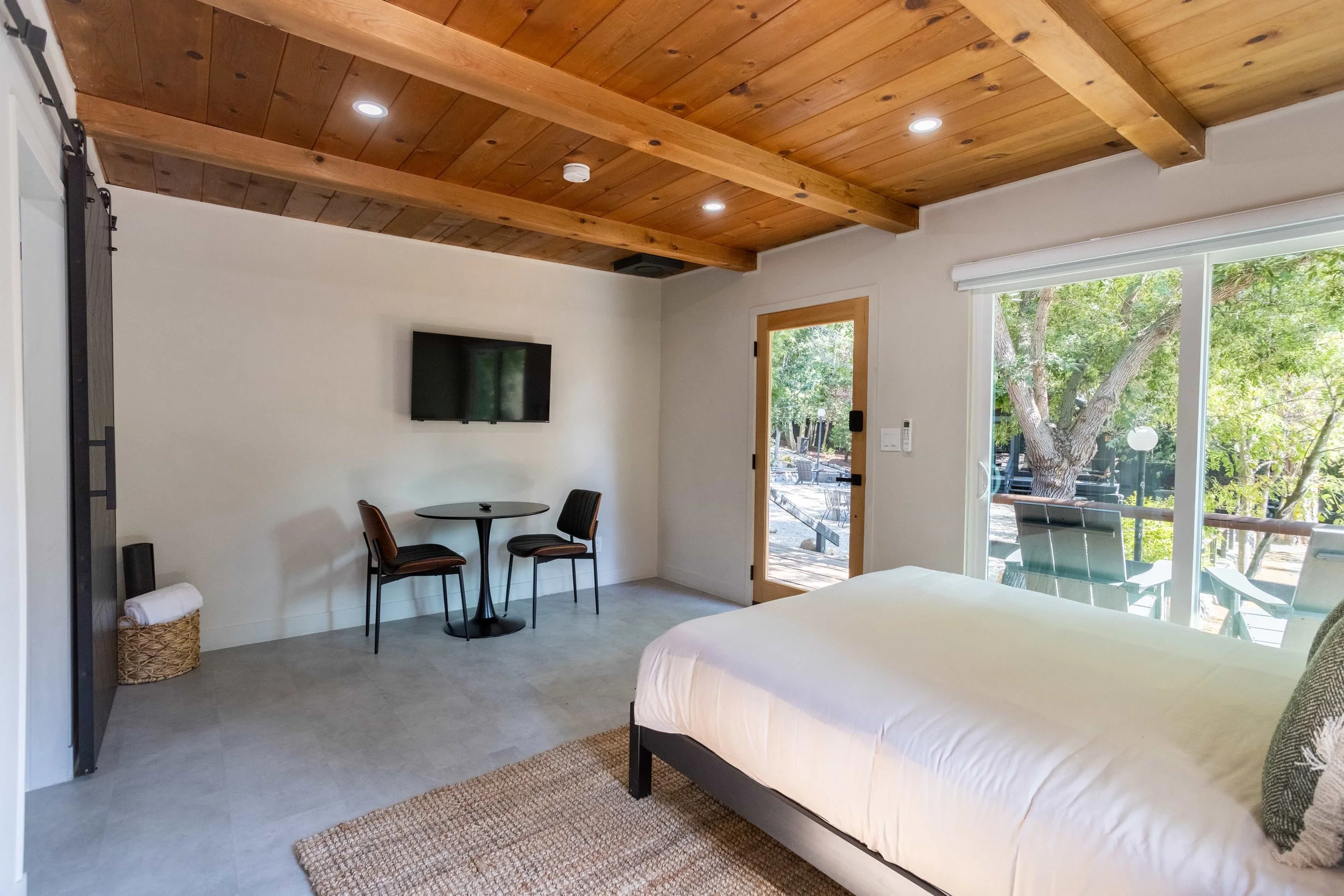 A bedroom with a wooden ceiling, a bed with white linens, a small round table with two black chairs, a wall-mounted TV, and a sliding glass door leading to an outdoor patio with trees and chairs.
