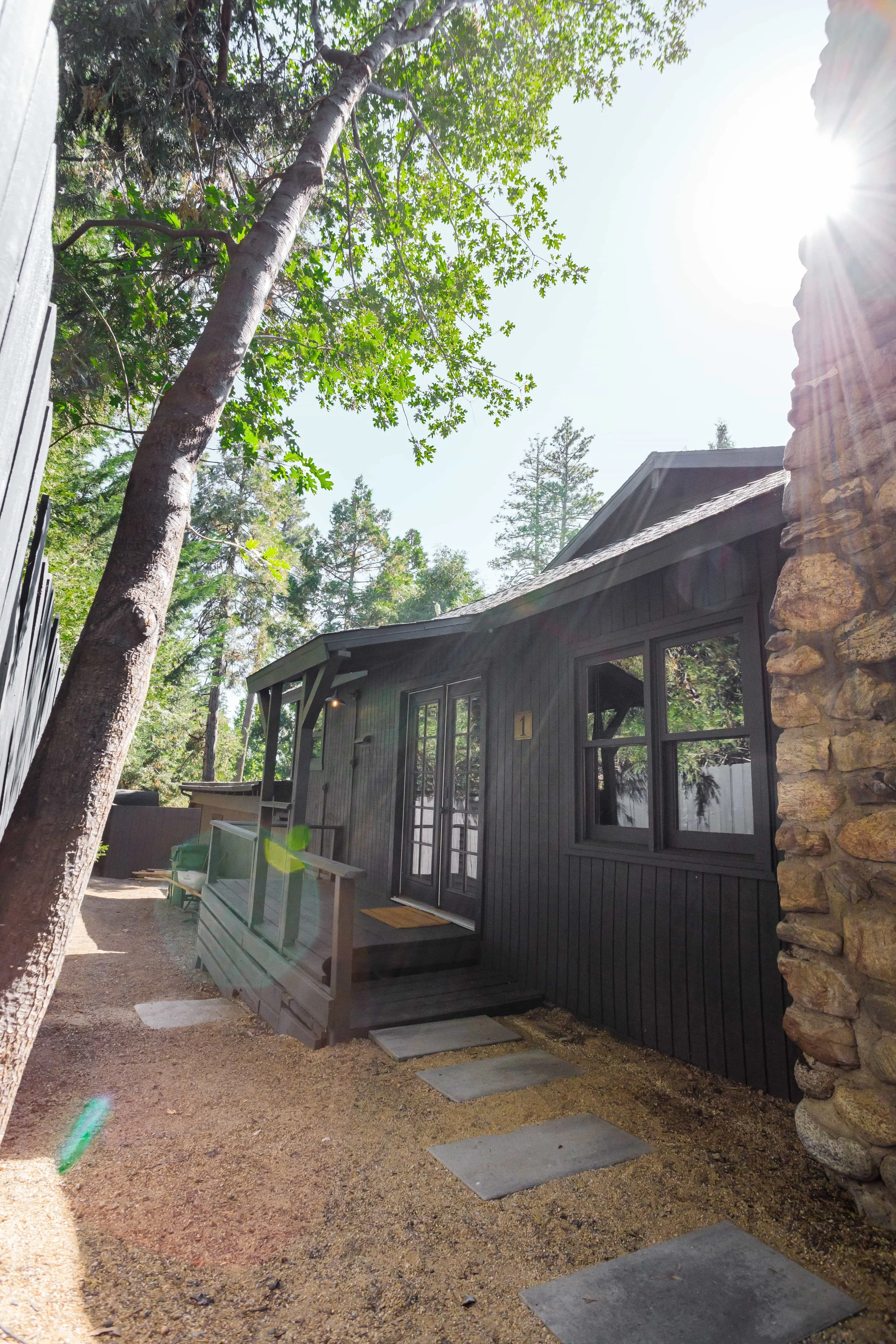 A small black wooden cabin with a stone chimney and front steps, surrounded by trees with sunlight filtering through.